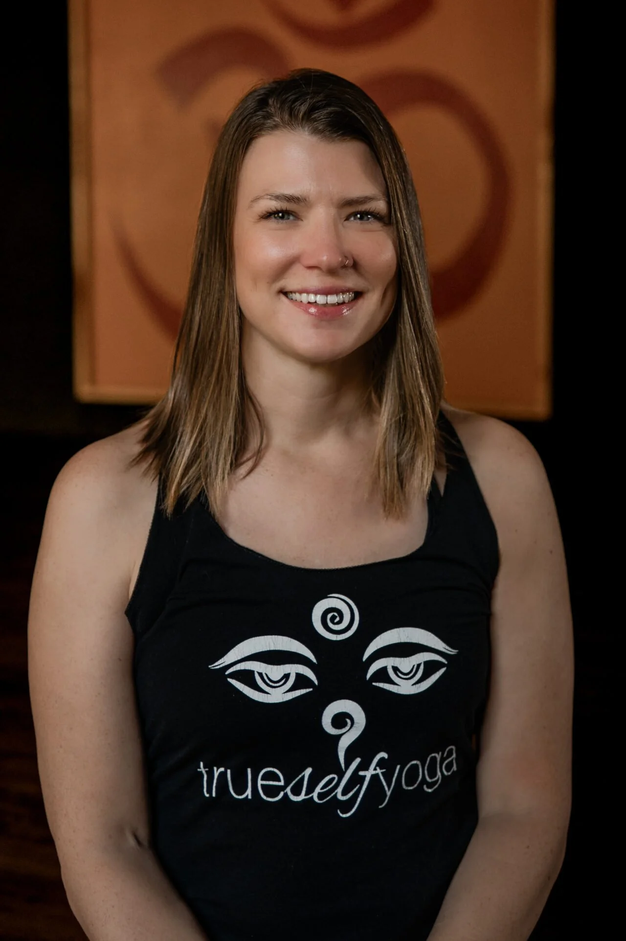 A woman with shoulder-length brown hair and a nose piercing, smiling, wearing a black tank top with a spiritual eye design and the words 'true self yoga' printed on it.
