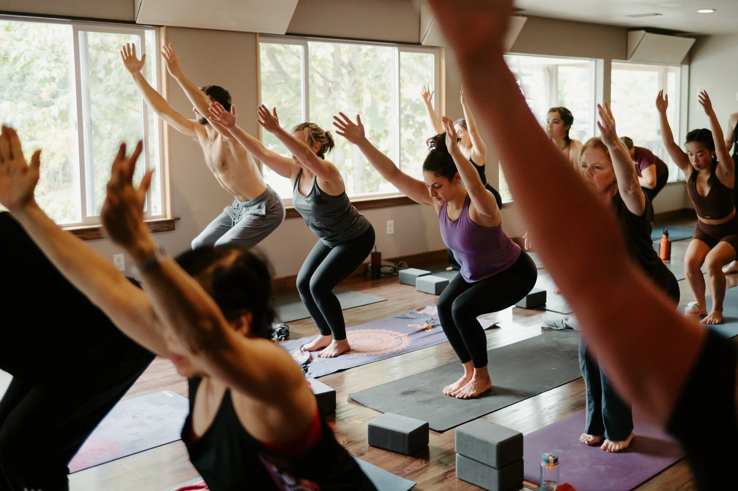 People participating in a group yoga or meditation class in a bright room with large windows, yoga mats, and blocks.