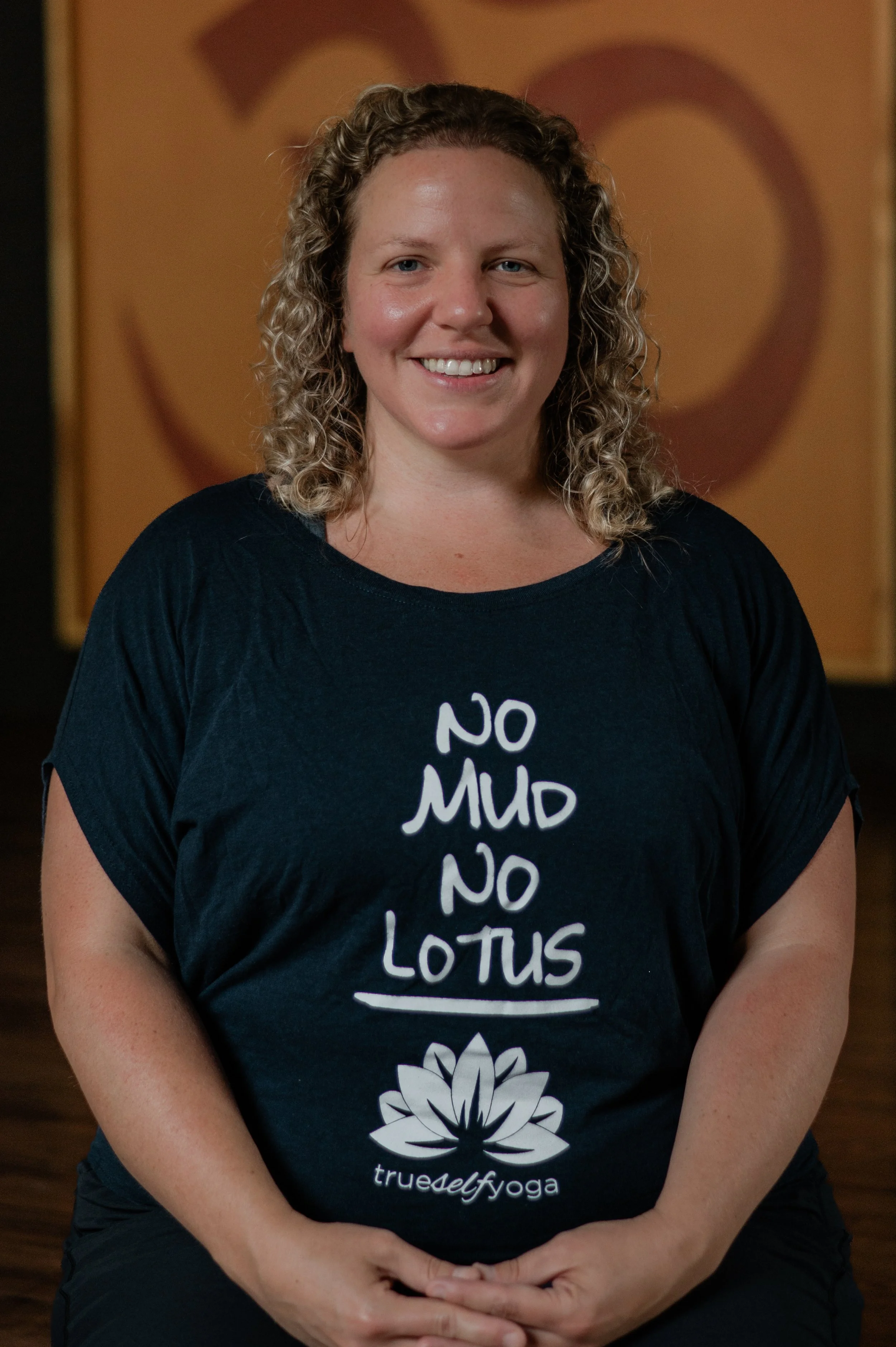 Woman with curly blonde hair smiling, wearing a dark T-shirt with a white lotus flower design and text that reads "truedef yoga." She is sitting in a yoga pose with her hands resting on her lap, in front of a wall with a large heart and circular design.