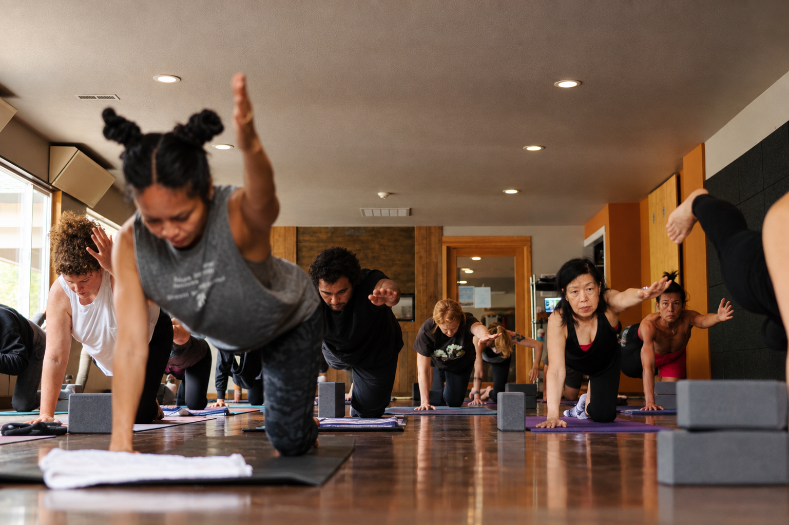 People participating in a yoga class, performing balancing poses on yoga mats in a studio with wooden walls.
