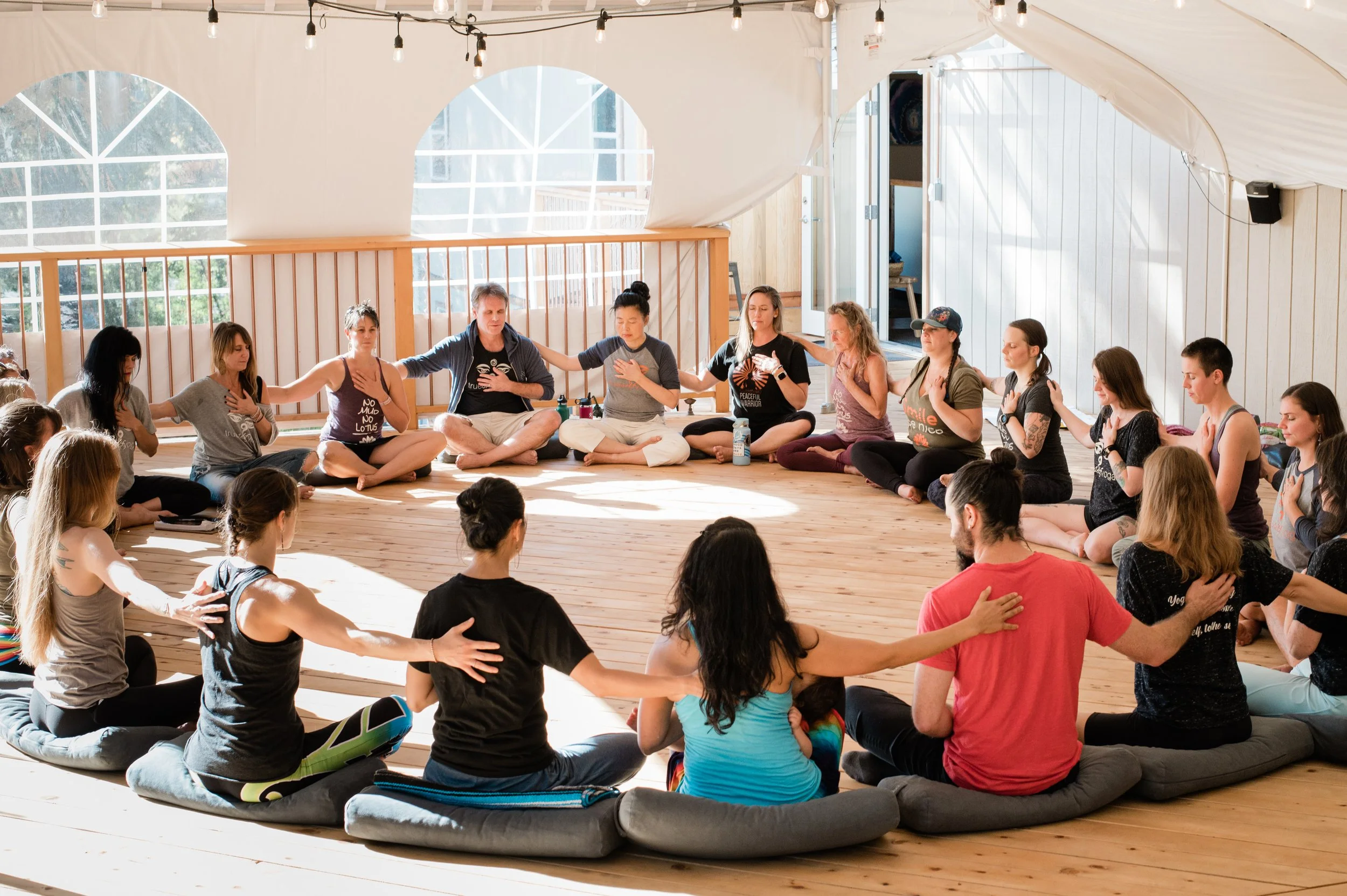 Group of people sitting in a circle in a well-lit indoor space, practicing meditation with hands over hearts and eyes closed.