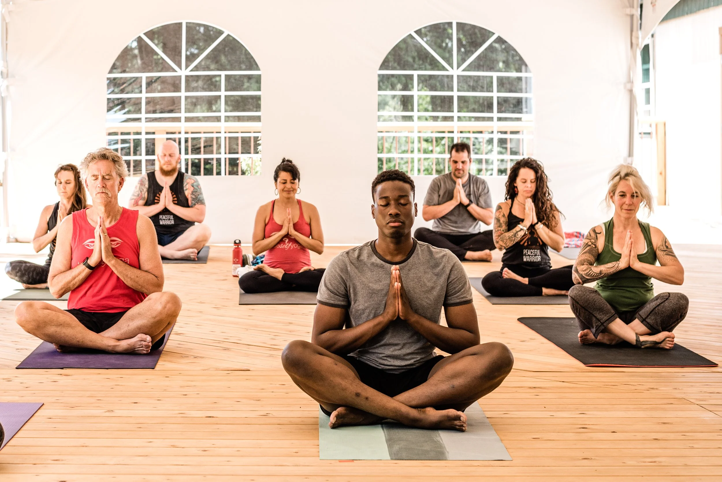 Group of people practicing yoga in a bright indoor studio with large arched windows, sitting cross-legged with hands in prayer position and eyes closed.