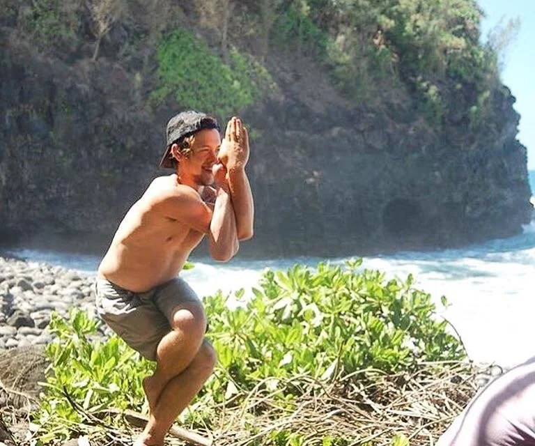 A shirtless man crouching on a rocky beach, smiling and holding his hands in a prayer position near his face, with greenery and ocean waves in the background.