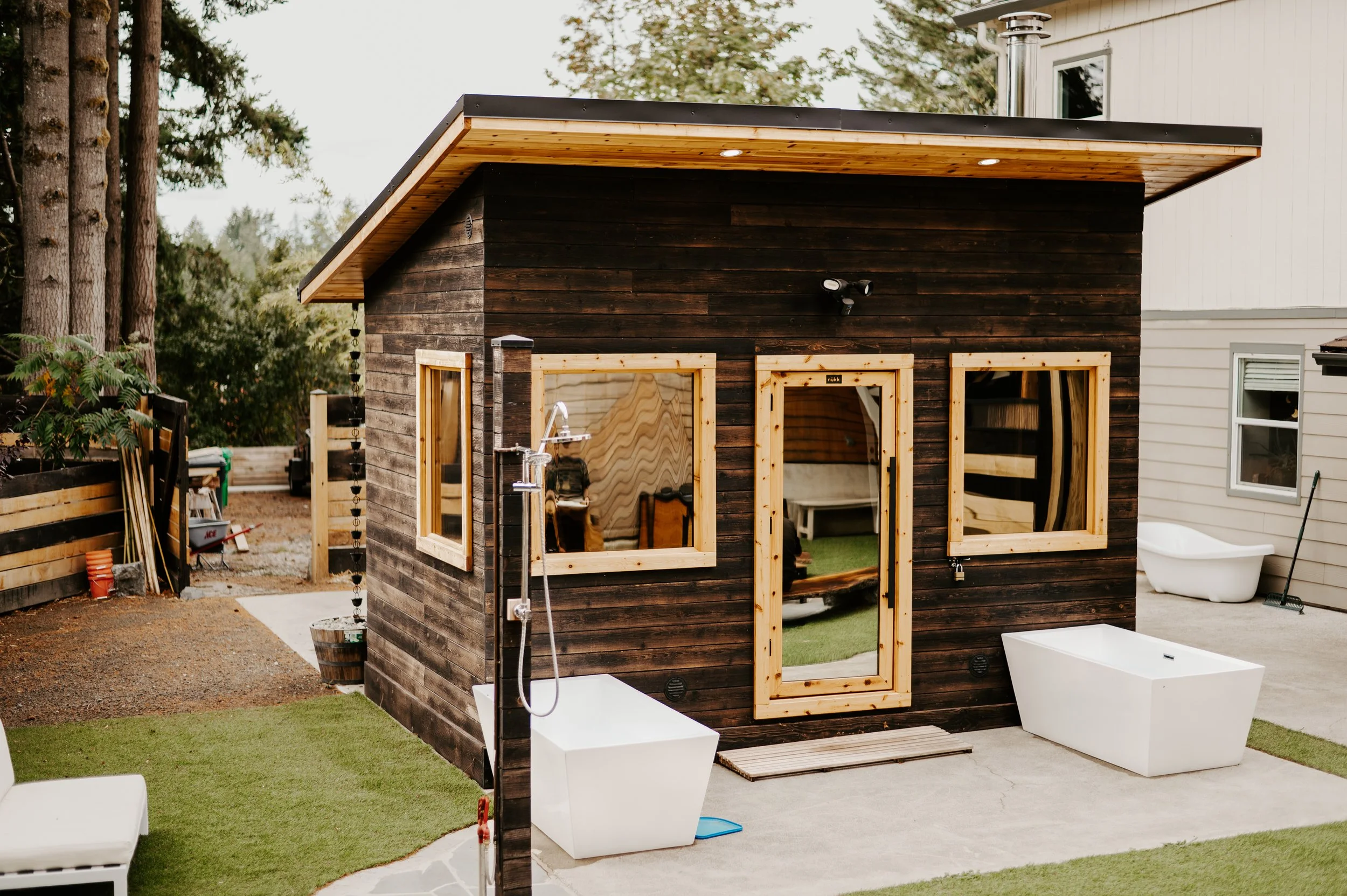 A small wooden backyard shed with large windows, a door, and a sloped roof, situated next to a white house, with outdoor furniture, a tub, and garden tools visible around it, surrounded by trees and a wooden fence.