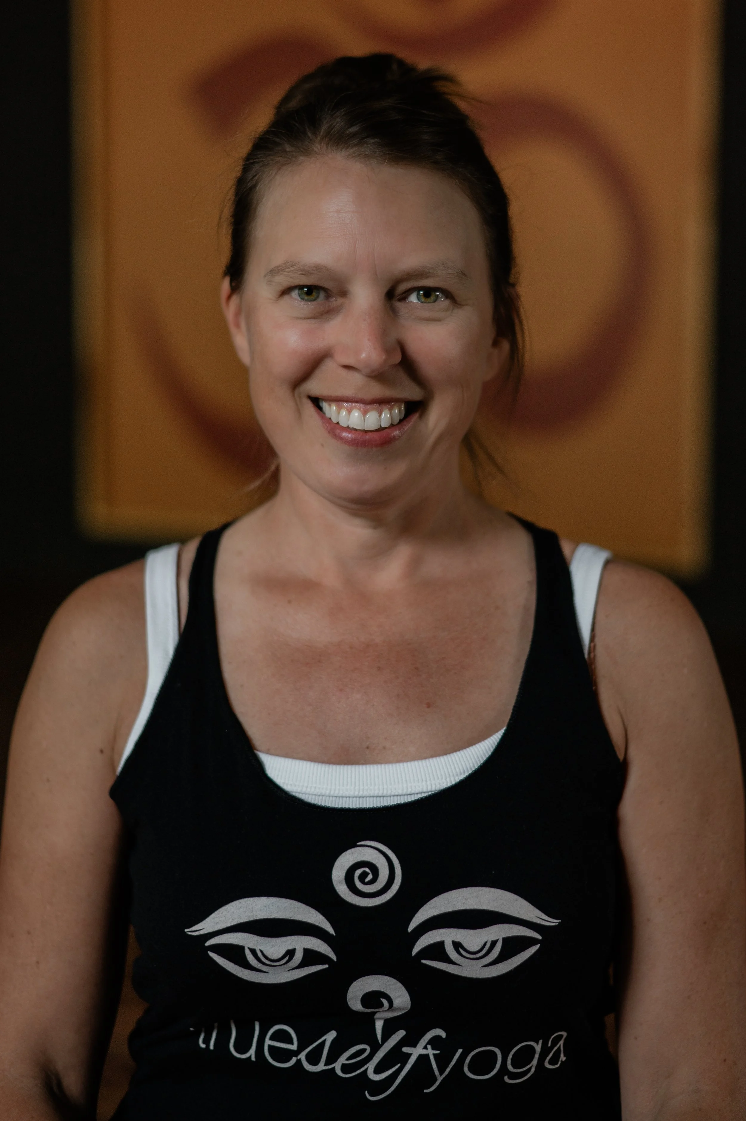 A woman smiling indoors with a background of abstract art featuring a spiral and eye design, wearing a black tank top with a white tribal face design and the words 'self yoga'.