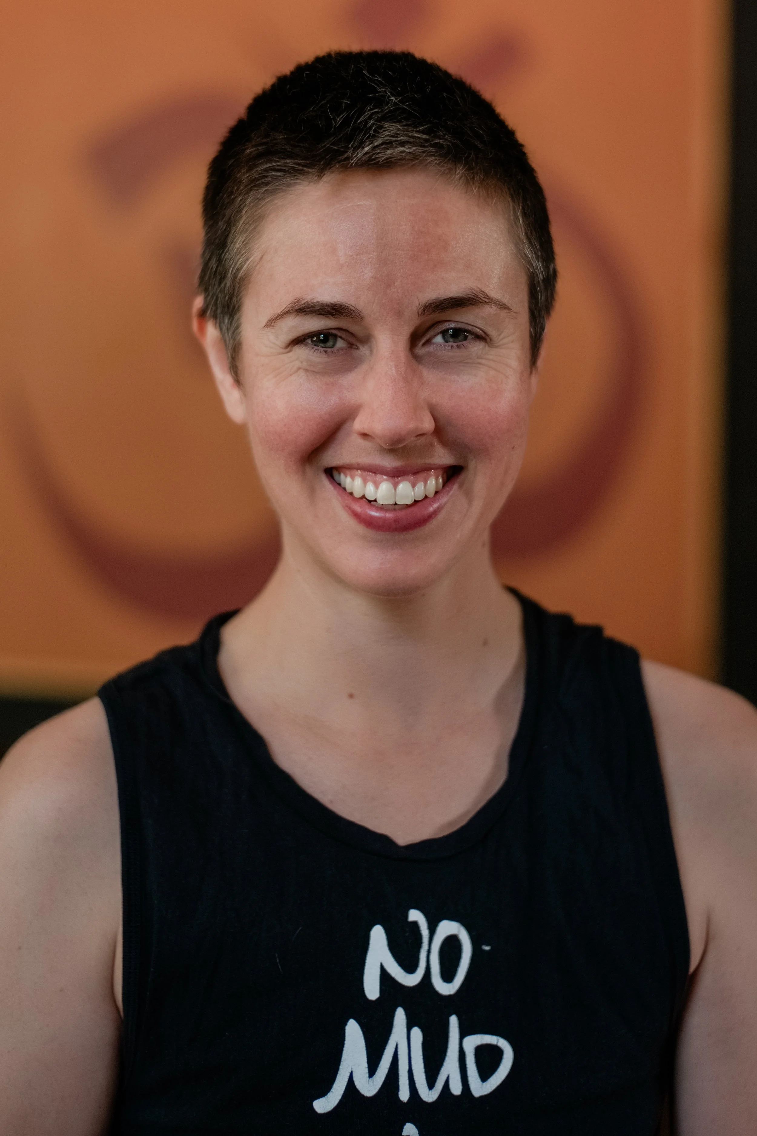 A woman smiling with short dark hair, wearing a black sleeveless top with white text, in front of a blurred orange background.