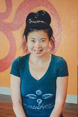A young woman with black hair styled in a high bun, smiling, wearing a blue t-shirt with a white design, standing in front of a pink and yellow patterned wall.