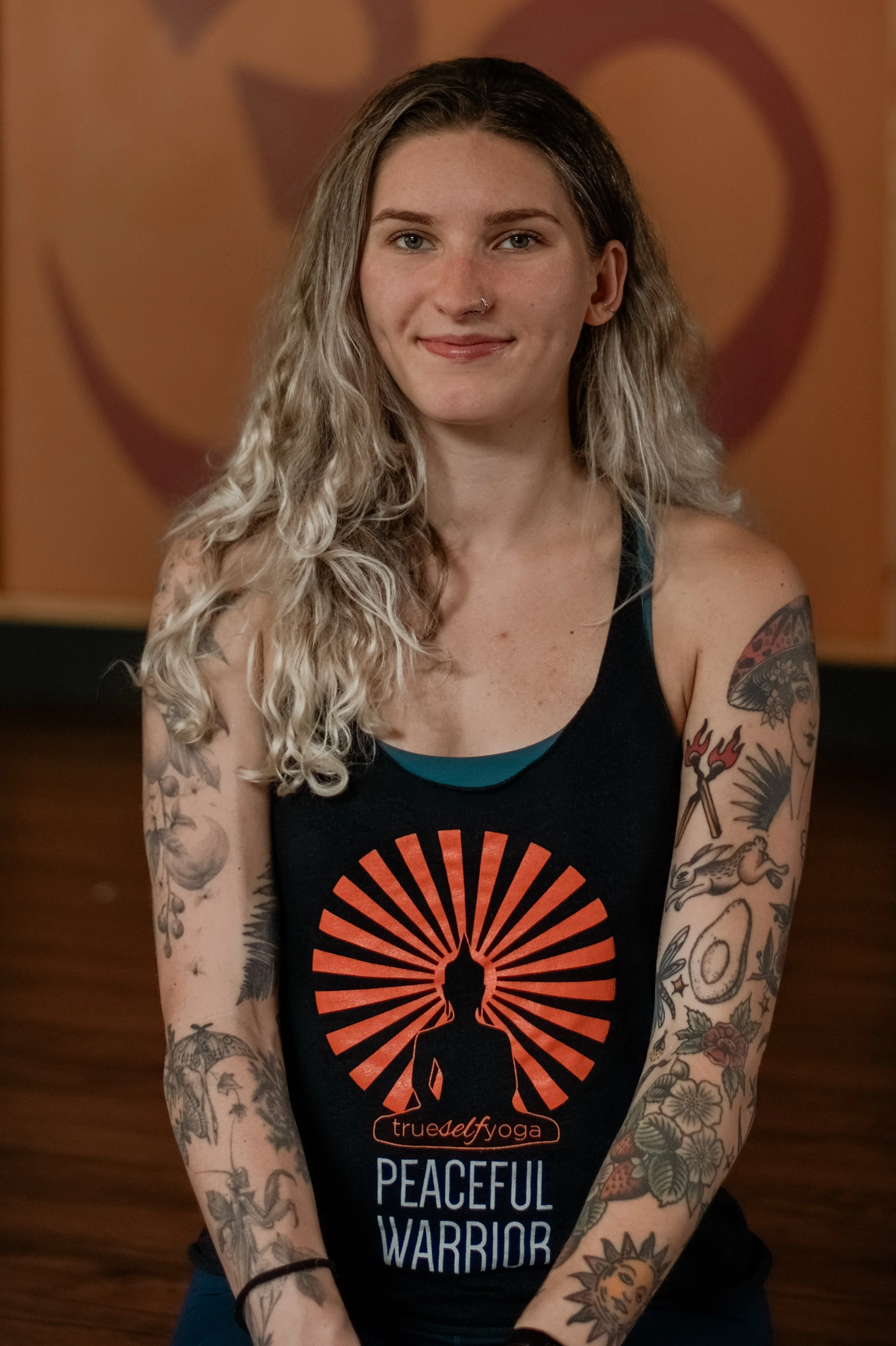 A young woman with long, wavy blonde hair, tattoos on both arms, and a nose ring, sitting and smiling at the camera. She wears a black tank top with a sun design and text that reads 'trueself yoga' and 'peaceful warrior.'