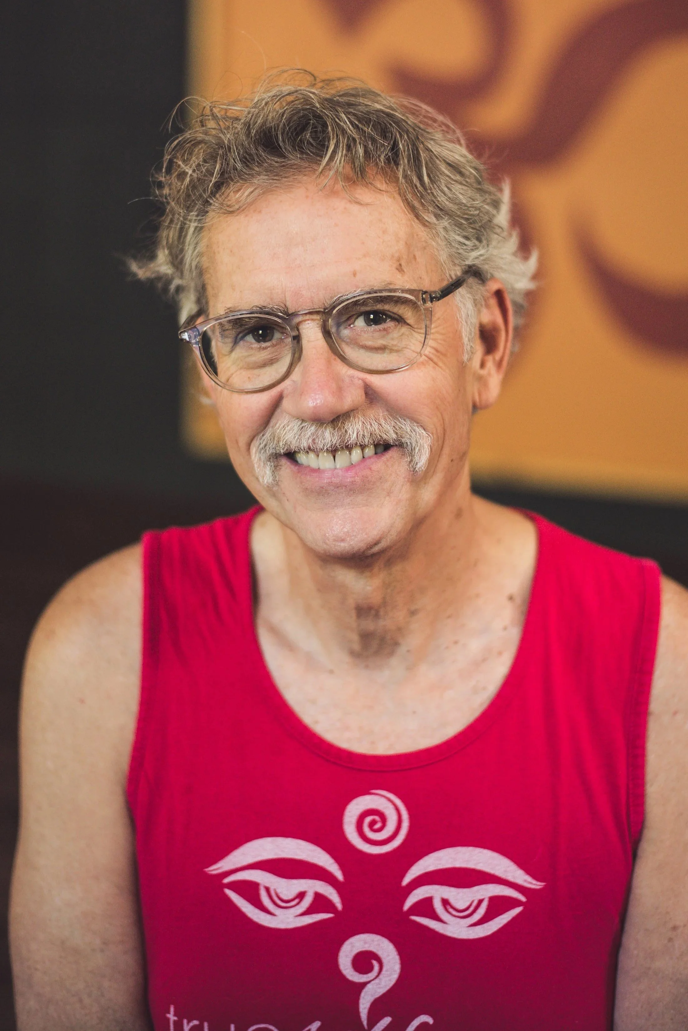 A smiling middle-aged man with gray hair, mustache, wearing glasses and a red sleeveless top with a white graphic design.