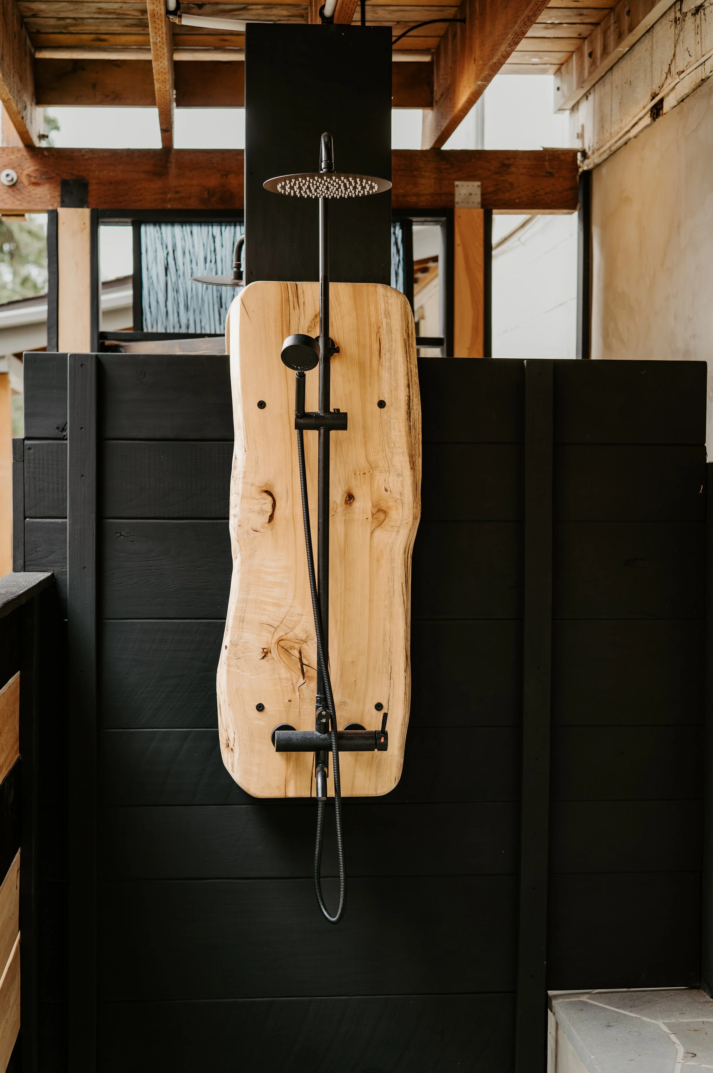 Wooden outdoor shower with black wall, mounted shower head, and handheld shower attachment, set against a black wall and wooden structure.