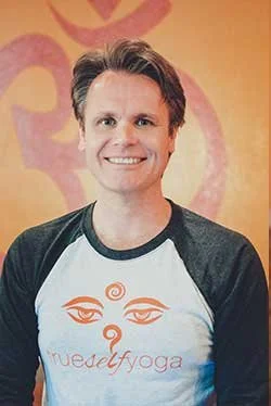 A smiling young man with short brown hair wearing a white and black t-shirt with an orange logo and text that says 'myself yoga' in front of a colorful, abstract background.