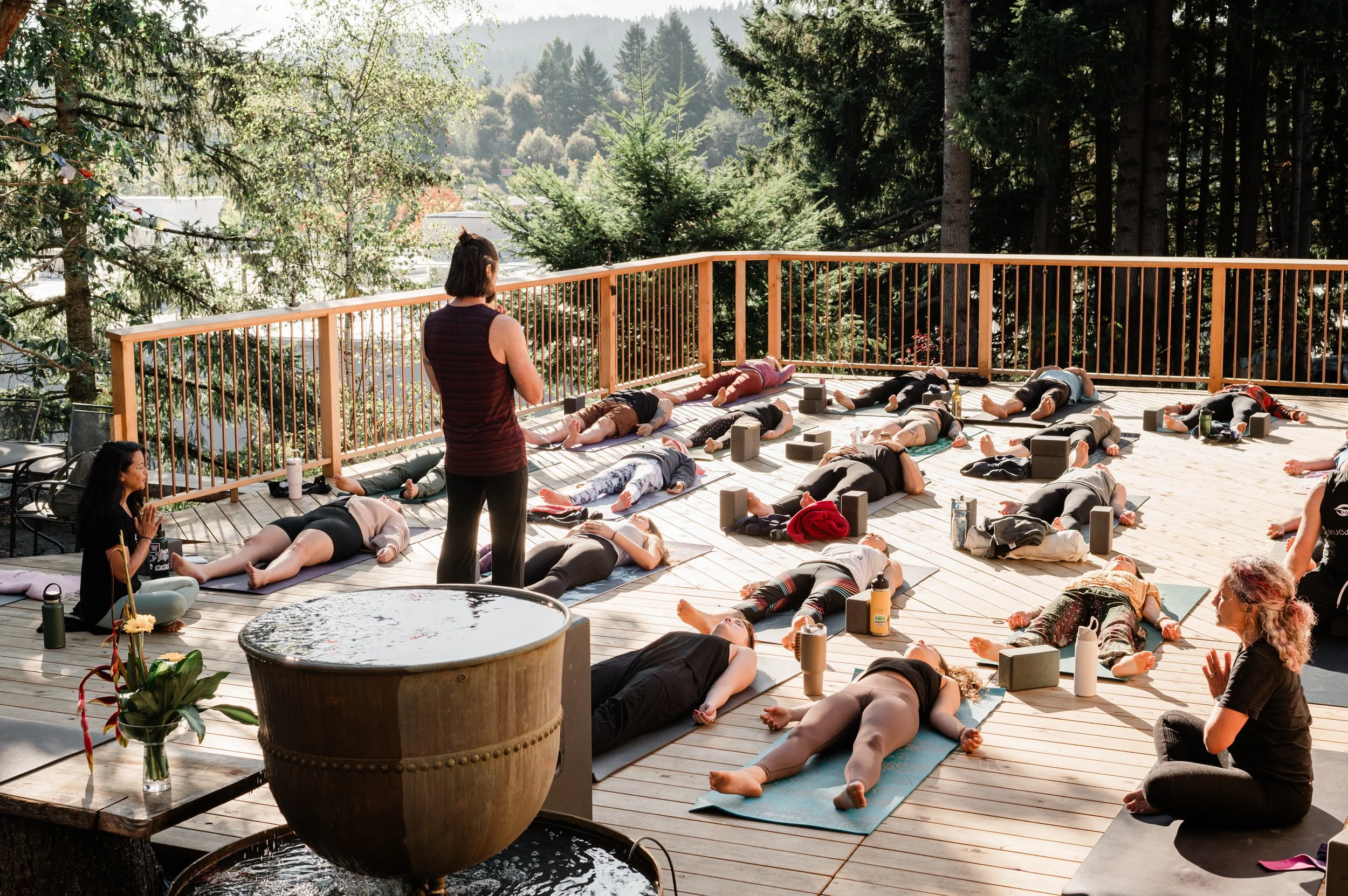 People participating in an outdoor yoga class on a wooden deck surrounded by trees, with some lying on yoga mats in a peaceful forest setting.