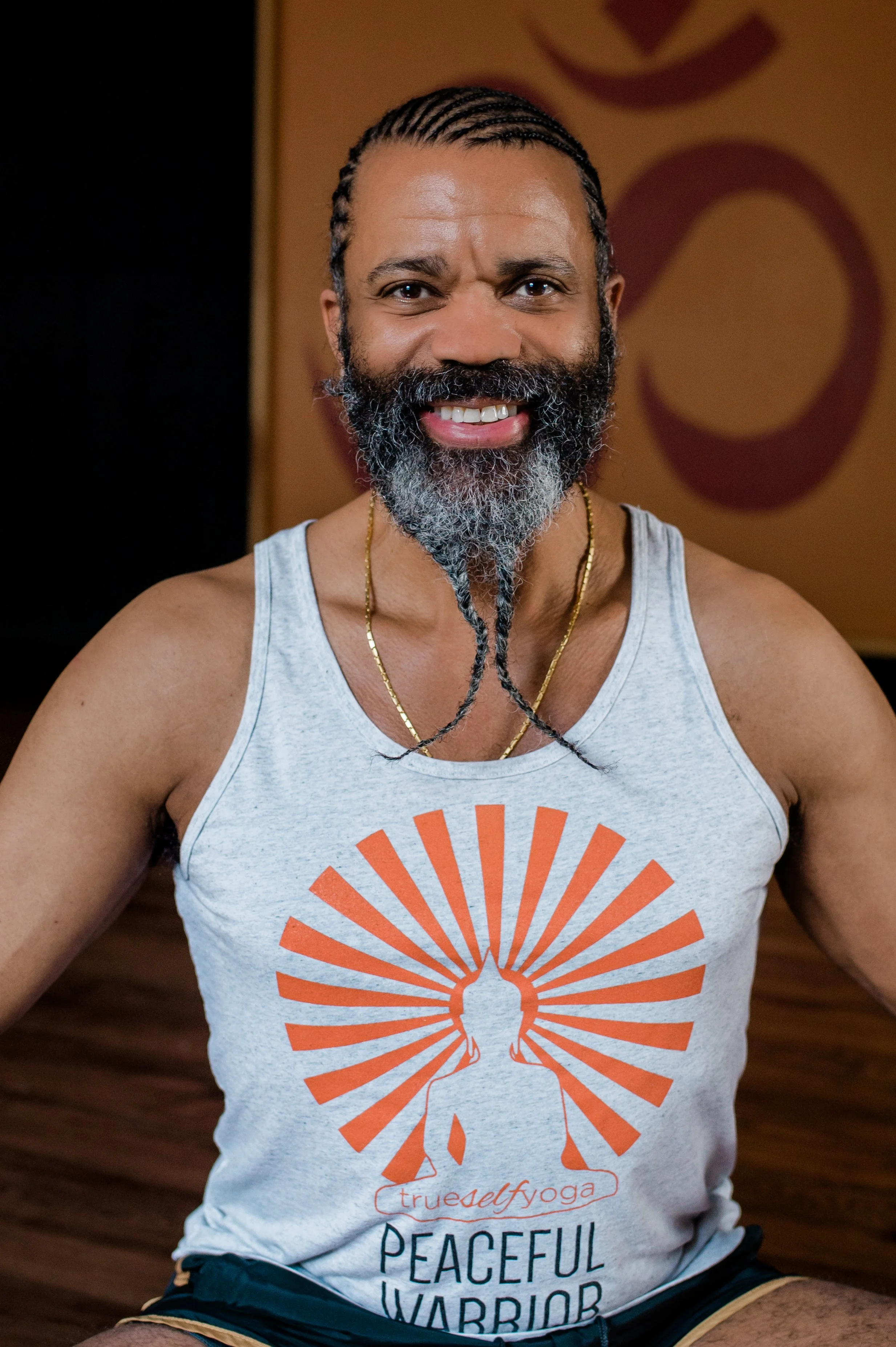 A man with a beard and braided hair, smiling, wearing a sleeveless shirt with a yoga logo and text about being a peaceful warrior, seated on the floor with a wooden background.