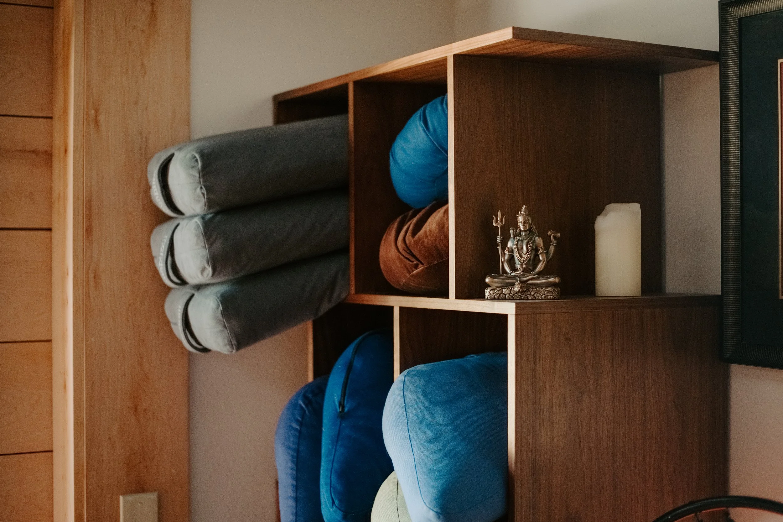 A wooden shelf unit with multiple compartments, holding rolled up blankets, a statue of a Hindu deity, and a candle, against a beige wall.
