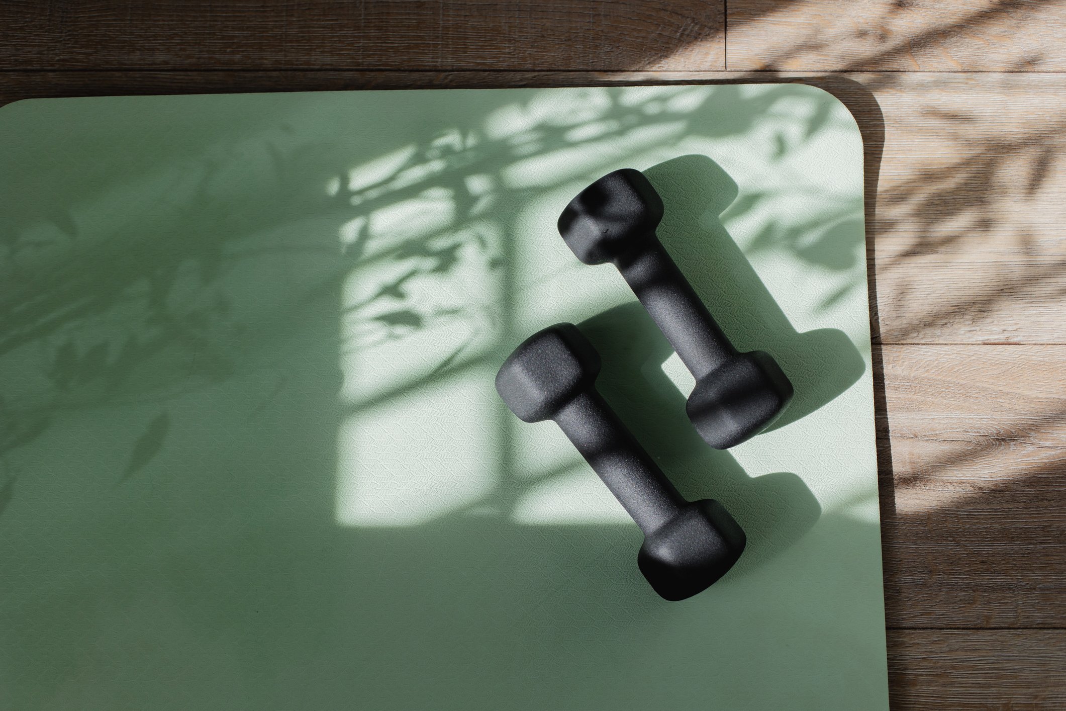 A pair of black dumbbells resting on a light green yoga mat with shadows of leaves, on a wooden floor.