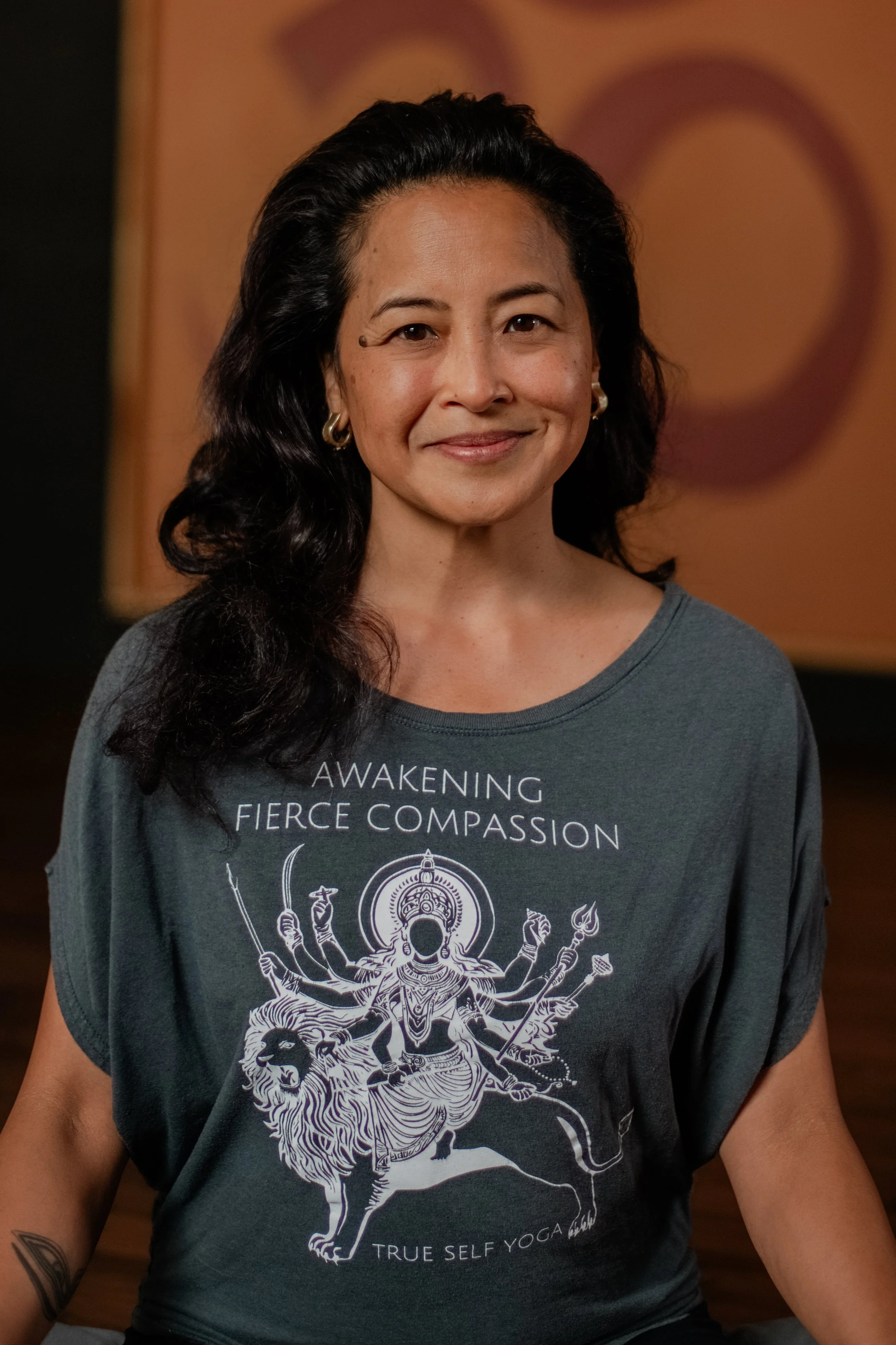 A woman with long black hair and hoop earrings is smiling at the camera, wearing a gray T-shirt with a Hindu goddess and lion illustration. The shirt has text that reads 'AWAKENING FIERCE COMPASSION TRUE SELF YOGA'.