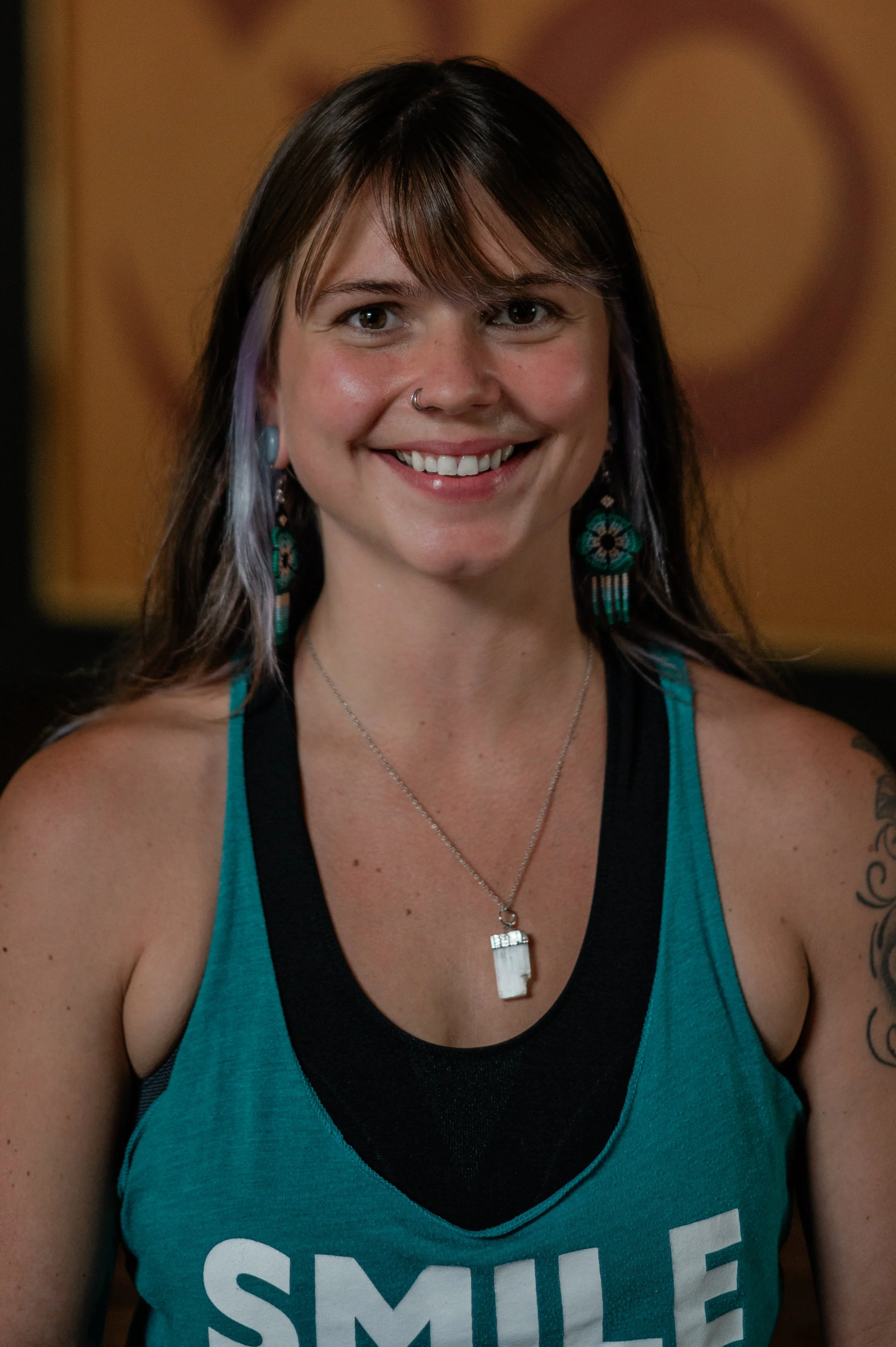 A woman with long dark hair, wearing a teal tank top with the word 'SMILE' printed on it, smiling and looking directly at the camera. She has a nose ring, colorful earrings, and is wearing a pendant necklace.