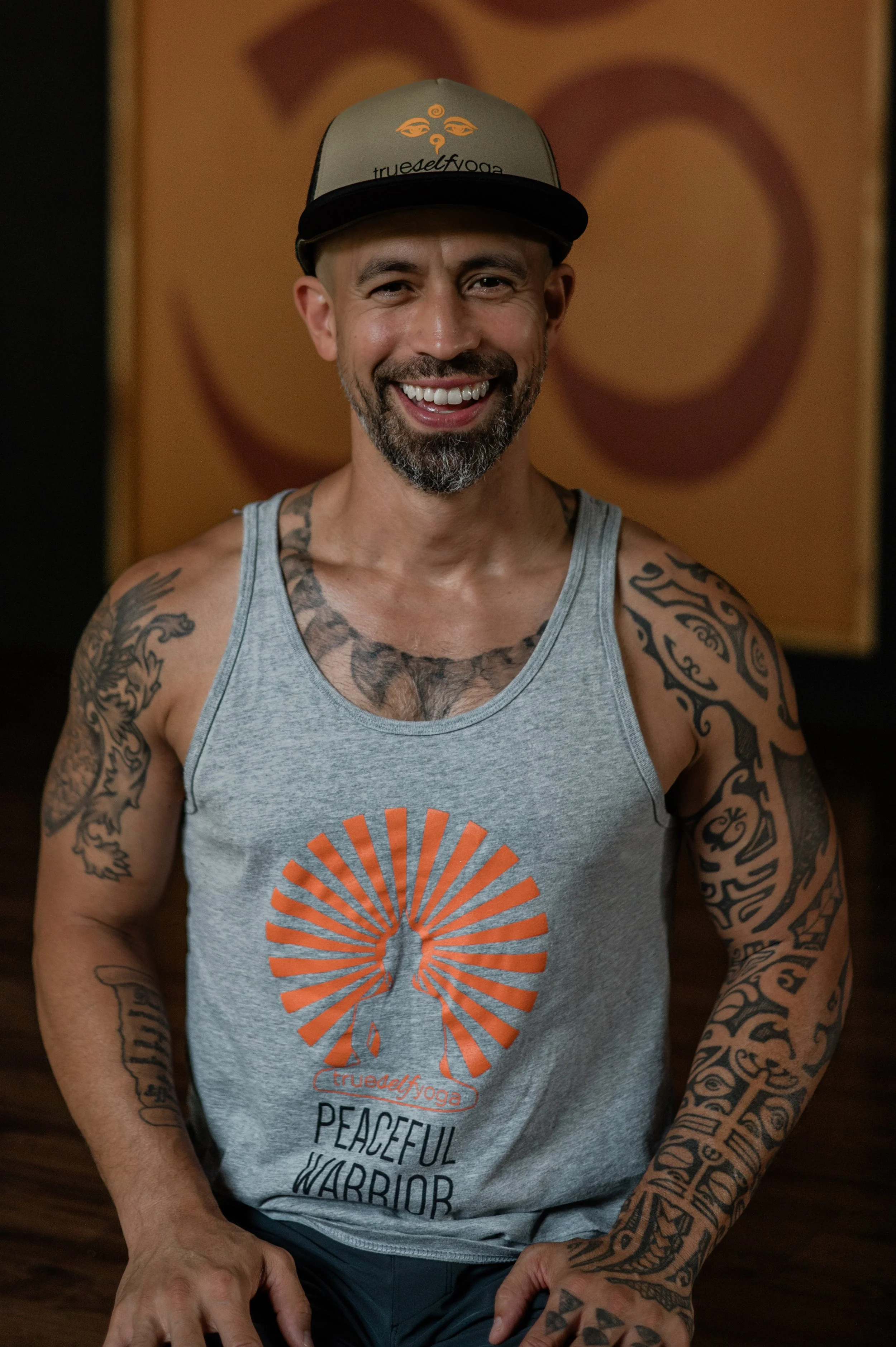 A smiling man with tattoos on his arms and chest, wearing a gray tank top with an orange logo and text, and a gray cap with text and design. He is standing in front of a background with warm-colored abstract art.