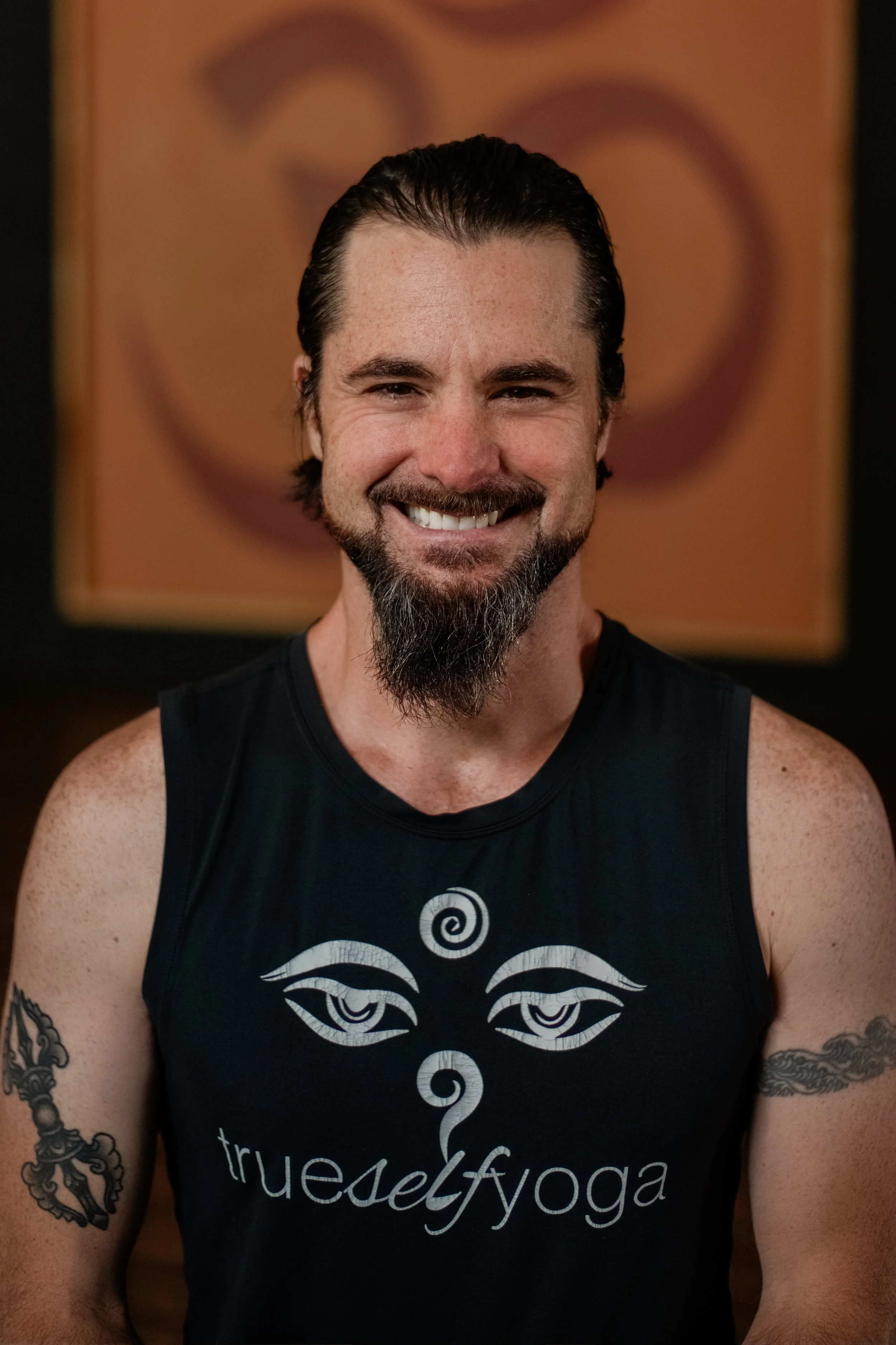 A man with dark, slicked-back hair and a beard, smiling, wearing a black tank top with a design and the words "true self yoga." He has tattoos on both arms. The background features an abstract artwork with swirling shapes and warm tones.