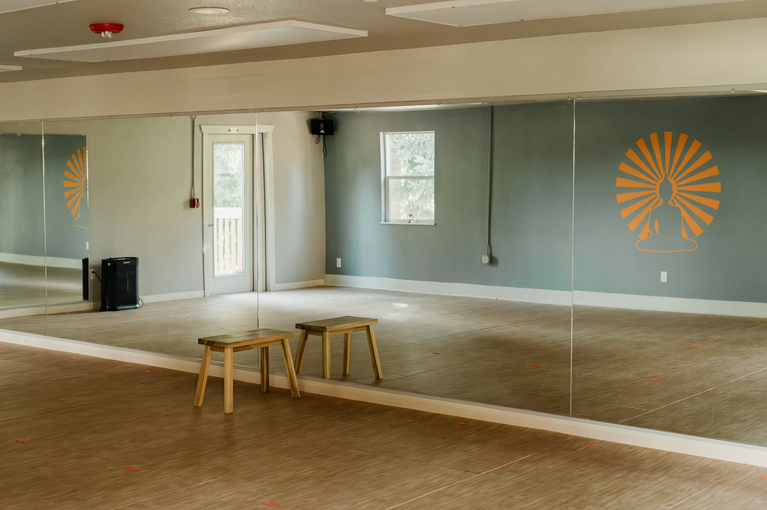 A dance studio with large mirrored walls, wooden floors, two small wooden benches, and a wall decoration of a meditating person with a sunflower behind their head, in an orange outline.
