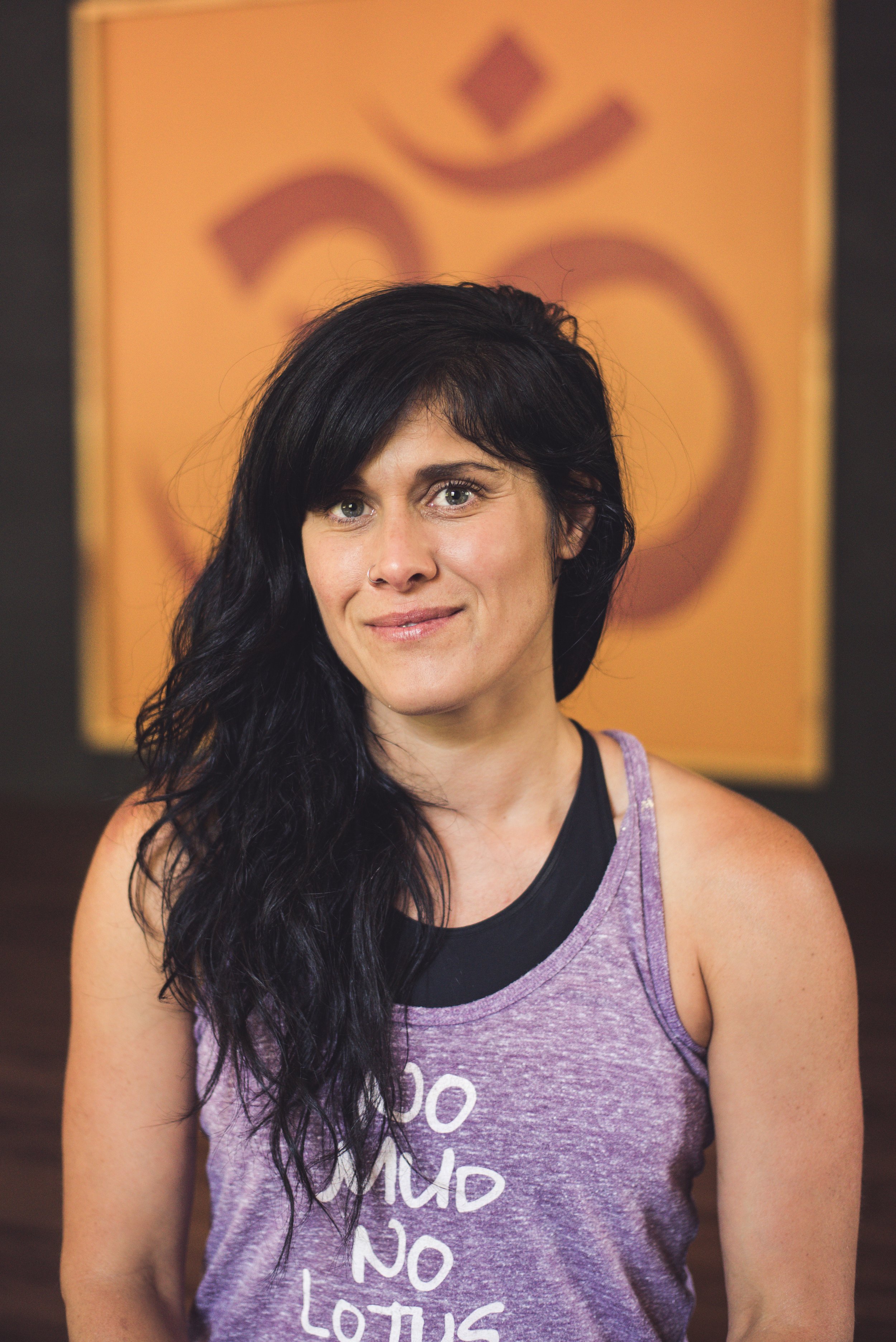 A woman with long black hair and a nose ring, wearing a purple tank top with white writing, smiling in front of a yellow-orange background with partially visible text.