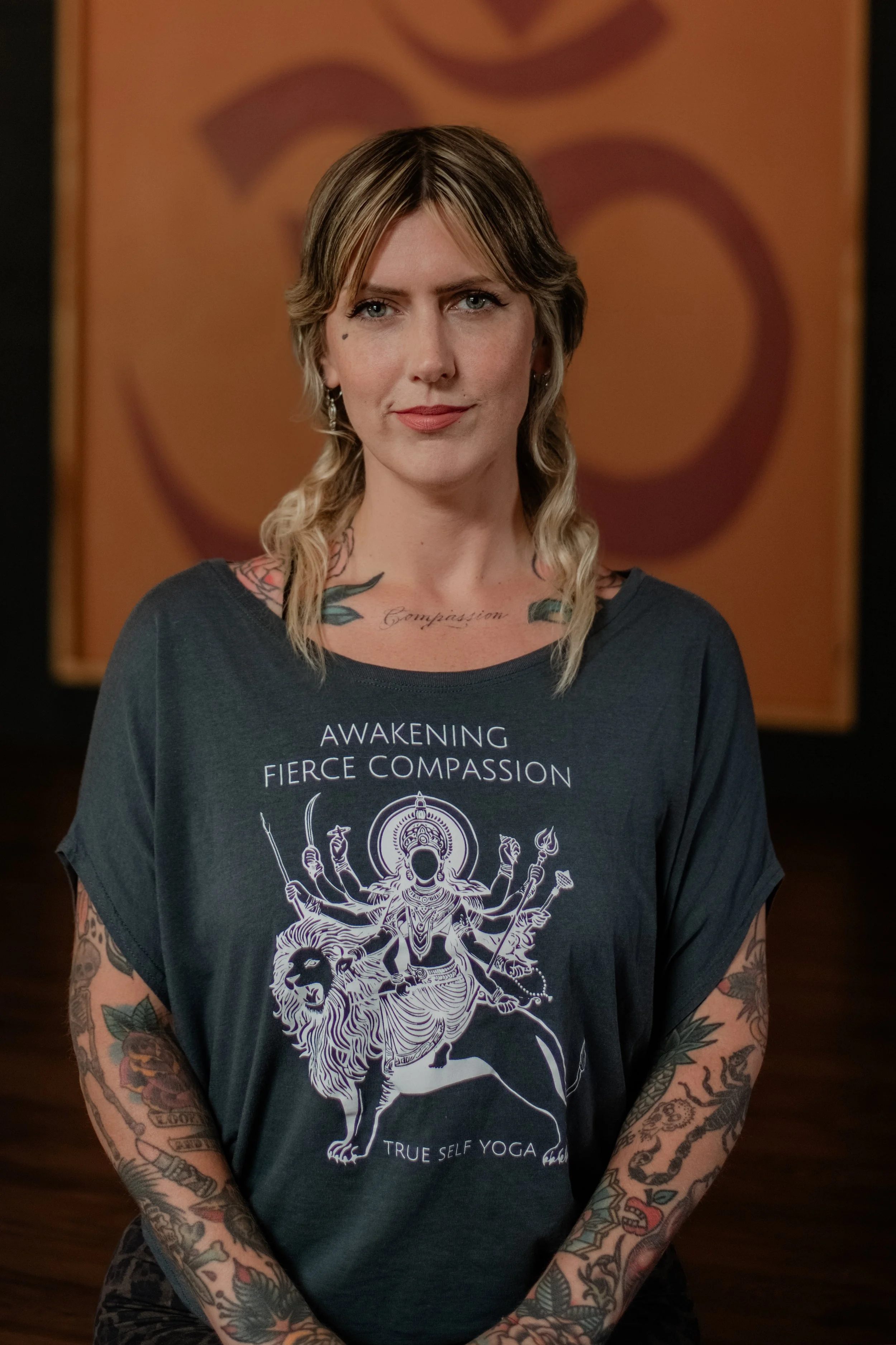 A woman with tattoos on her arms and neck, wearing a black T-shirt with white graphic and text, standing in front of a background featuring a large orange and brown artwork.