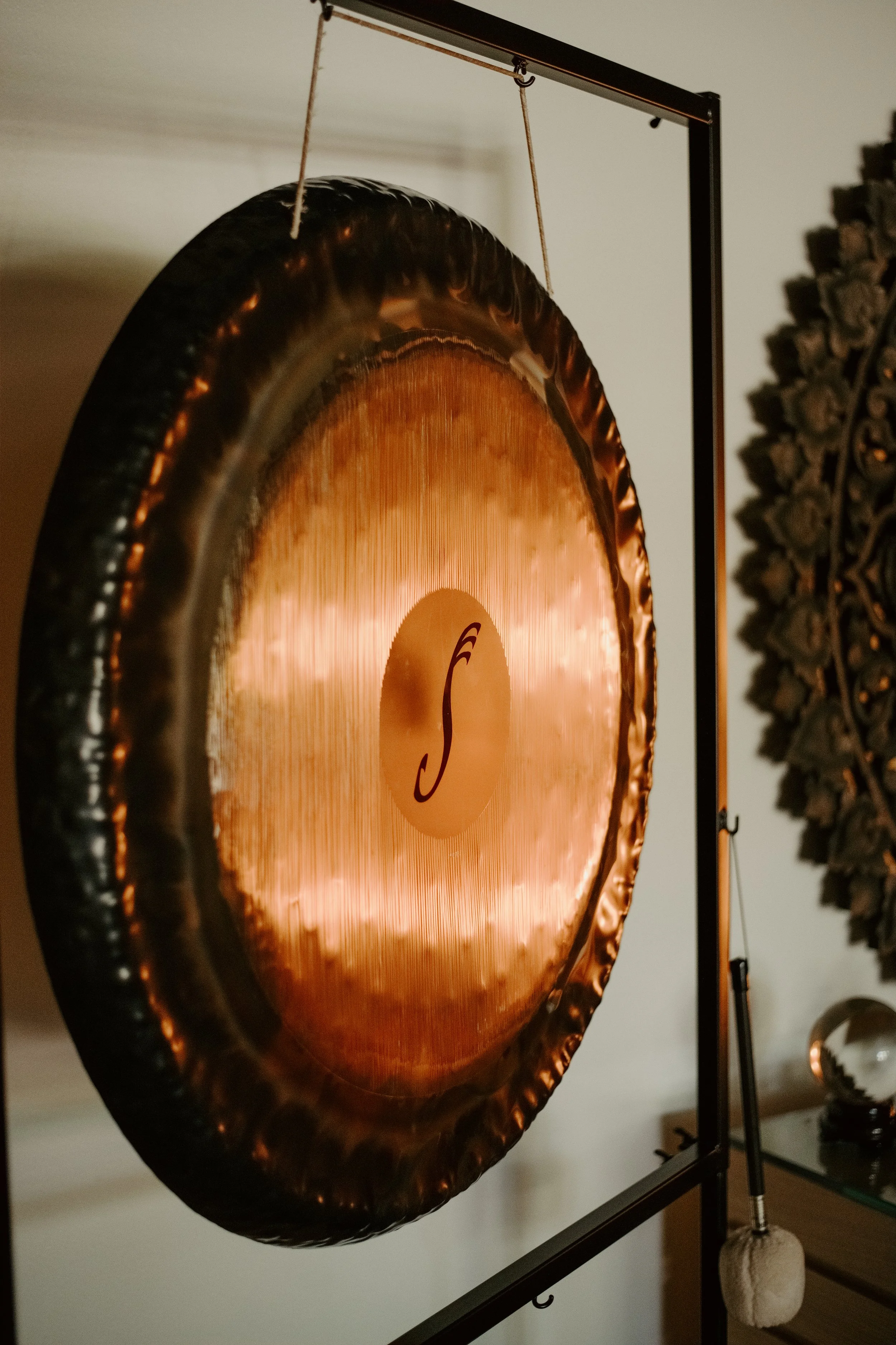 A large copper gong with a circular logo in the center, mounted on a black metal stand. The logo has a stylized S letter. The background shows a wall and some decorative elements.