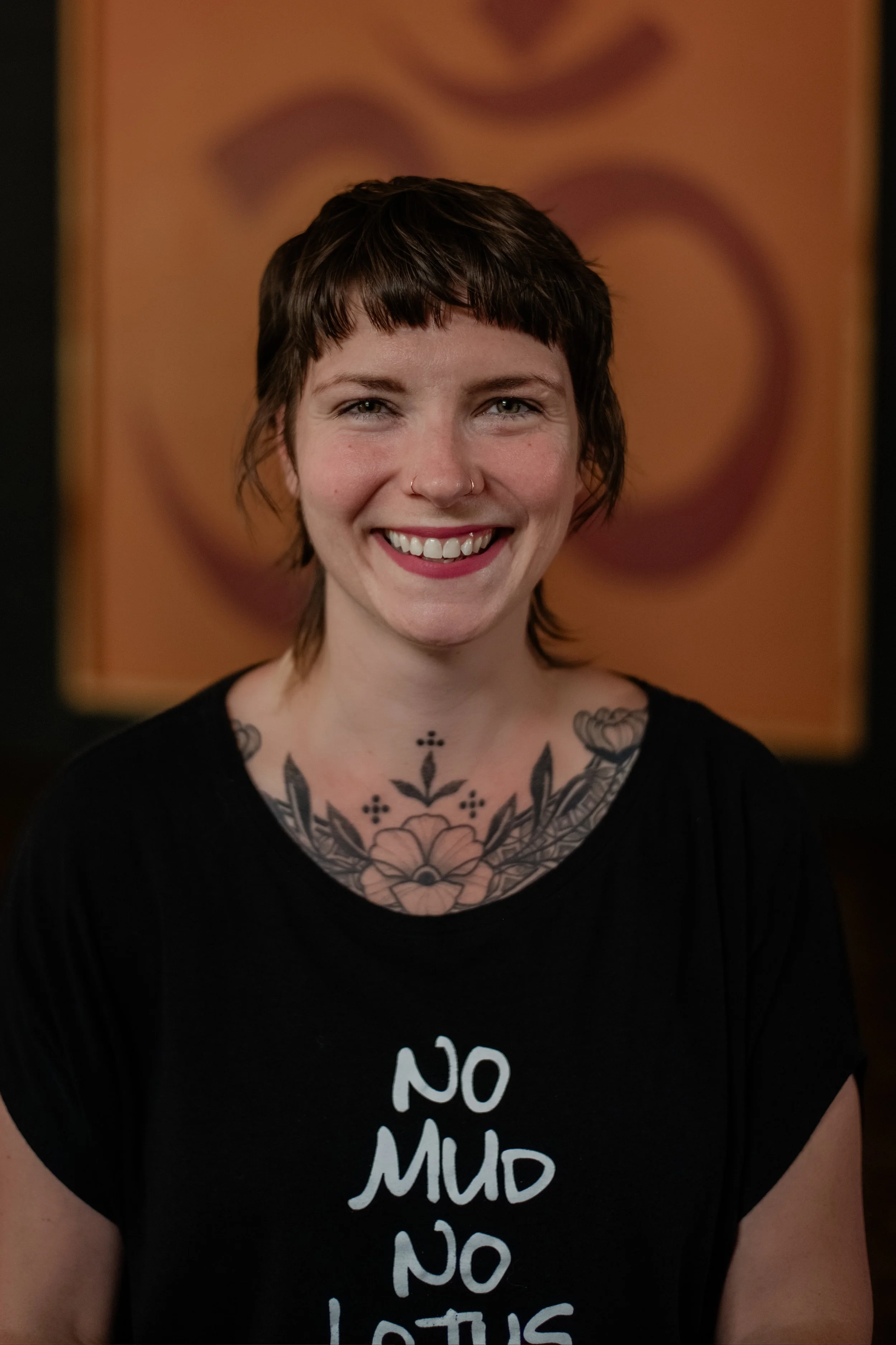 A smiling woman with short brown hair, tattoos on her chest, wearing a black shirt with white text, standing in front of an orange background with circular patterns.