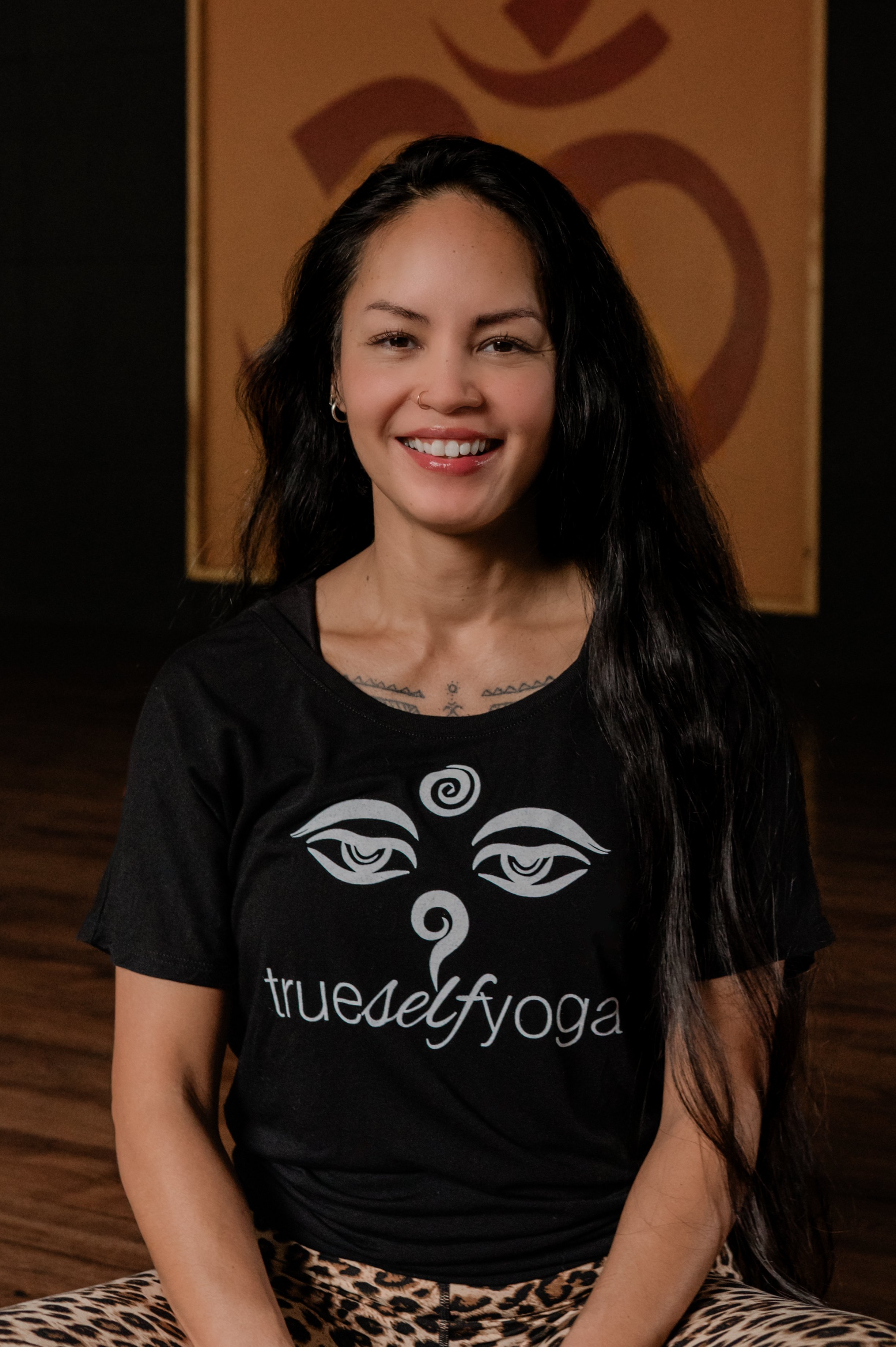 A woman with long dark hair, wearing a black t-shirt with a design and the words "trueself yoga" on it, sitting cross-legged in front of a wooden wall and a backdrop with a large heart and abstract art.