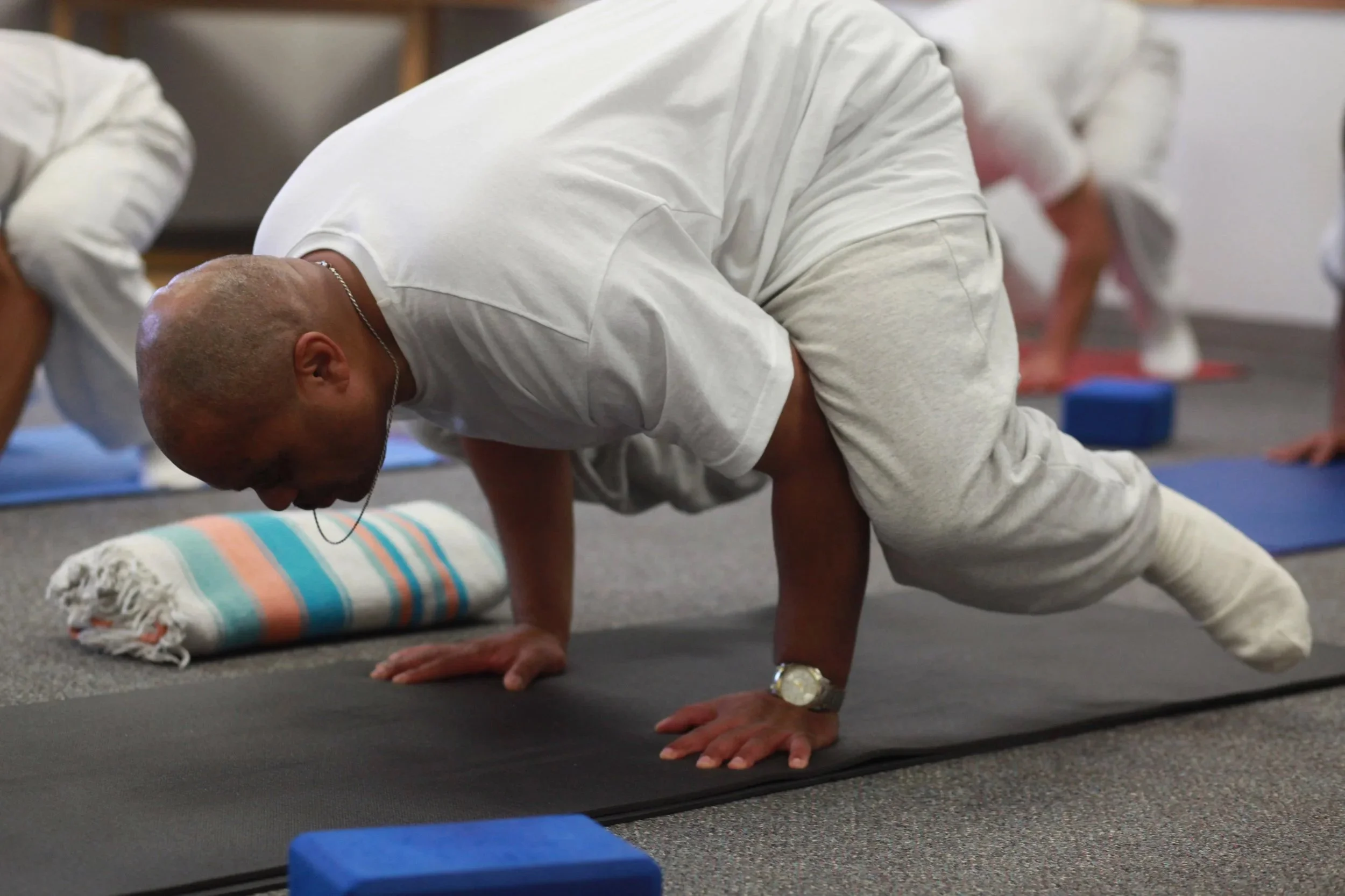 A man wearing a white shirt and beige shorts performing a plank yoga pose on a black mat, with a rolled towel nearby, in a room with several other people doing similar poses.