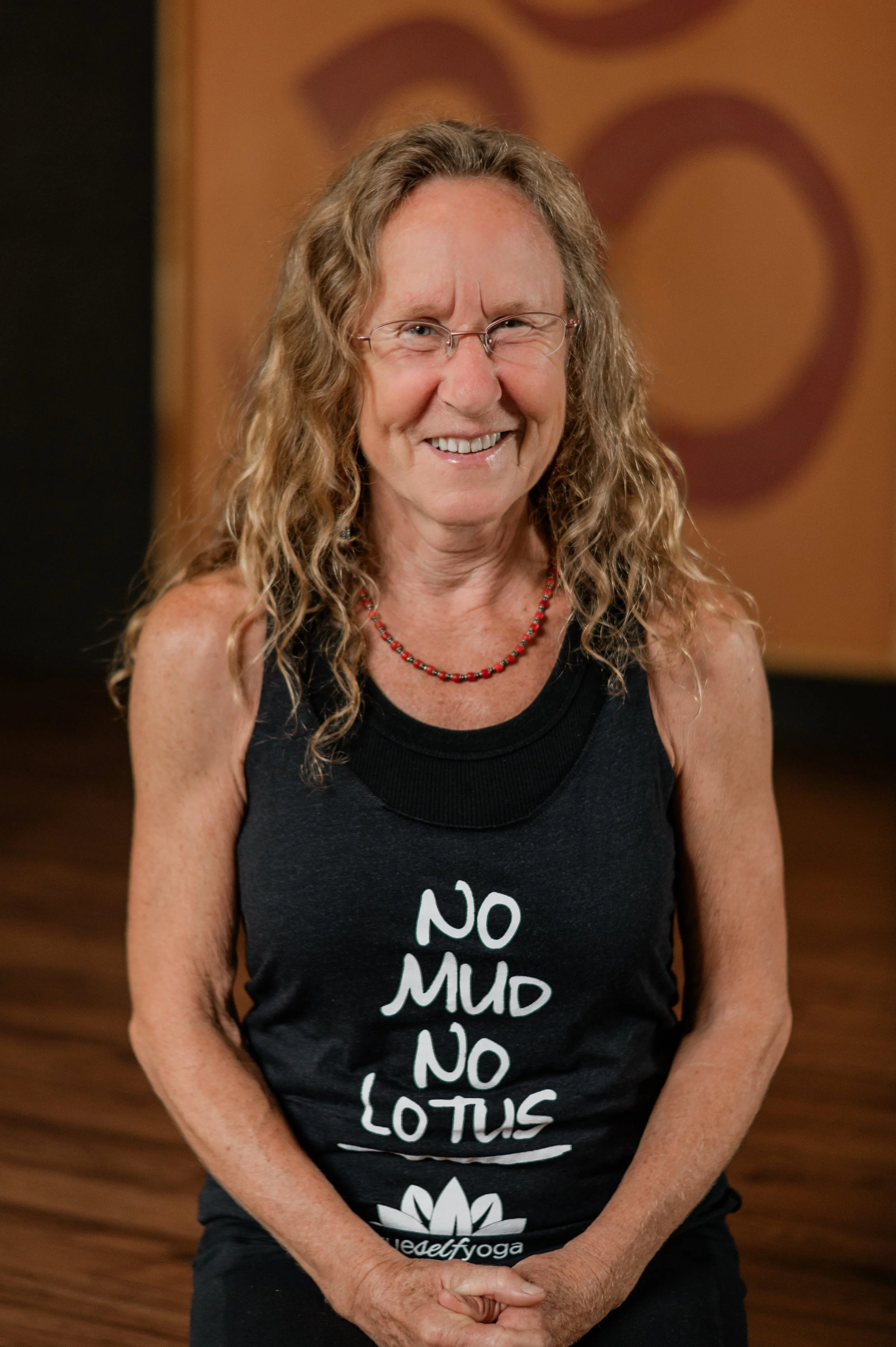 A smiling woman with curly hair and glasses, wearing a black tank top with the words 'NO MUD, NO LOTUS' and a lotus flower graphic, in a room with wooden flooring and a brown background.