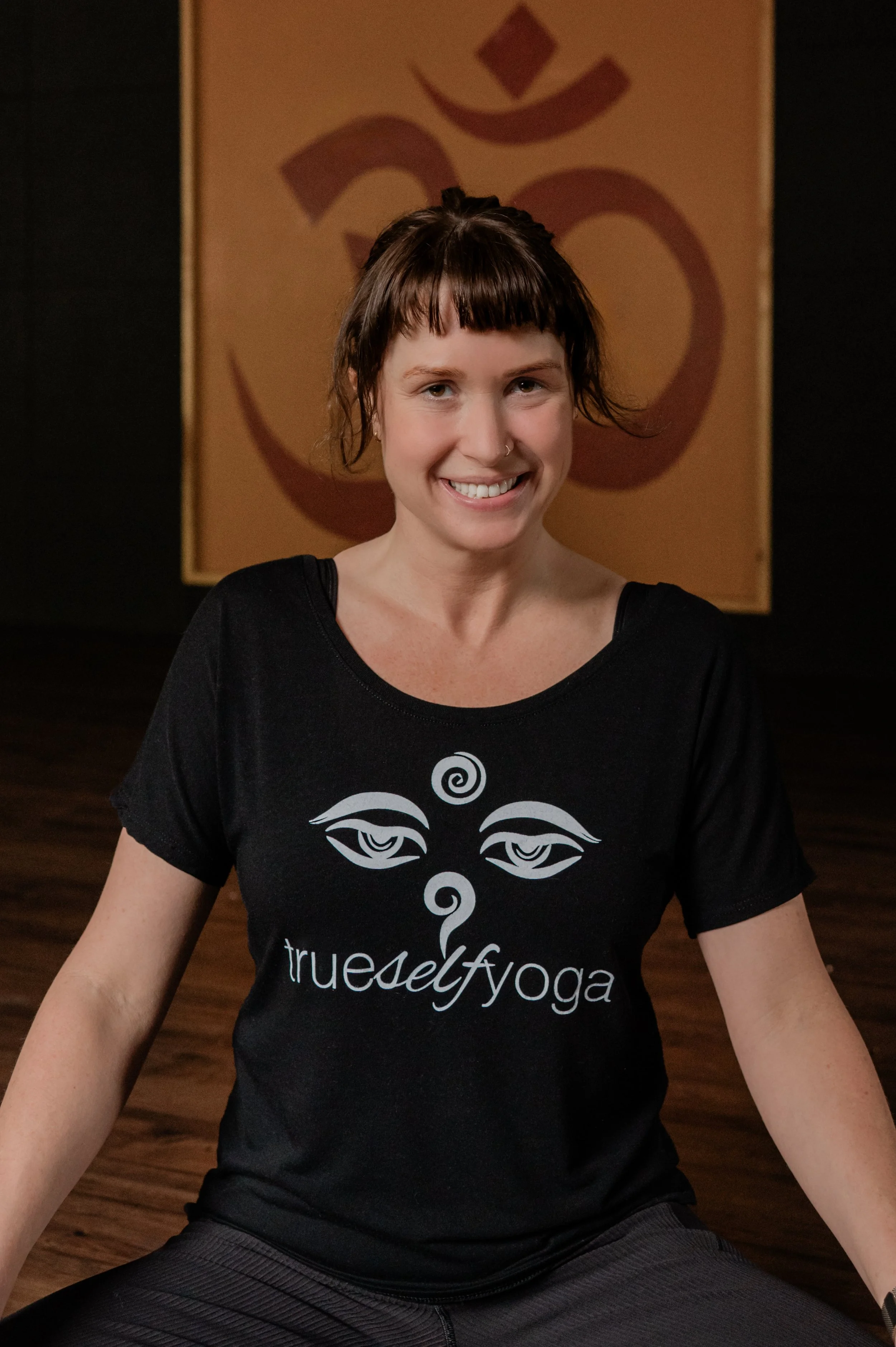 Woman practicing yoga in a studio, sitting cross-legged with her hands on her knees, wearing a black 'true self yoga' t-shirt. A large Om symbol hangs on the wall behind her.