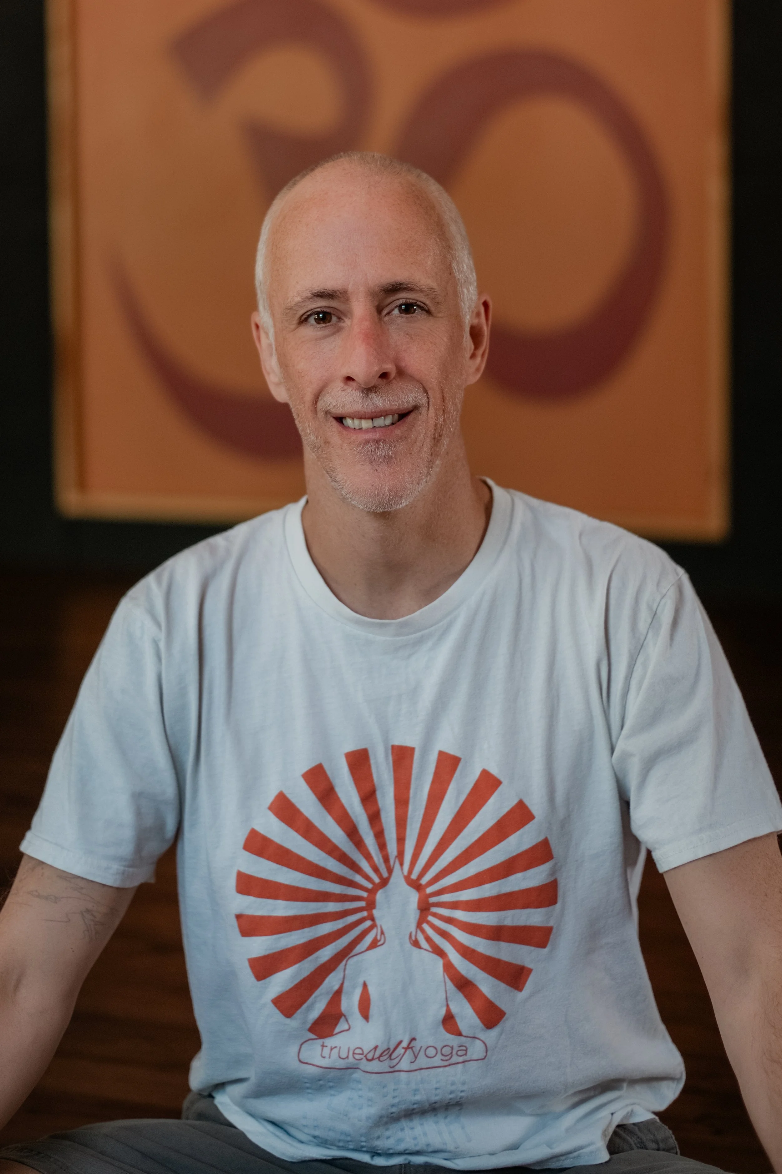A smiling man with a shaved head and gray beard, wearing a white T-shirt with an orange sun design and the words 'trueself yoga,' sitting in front of abstract wall art with warm tones.