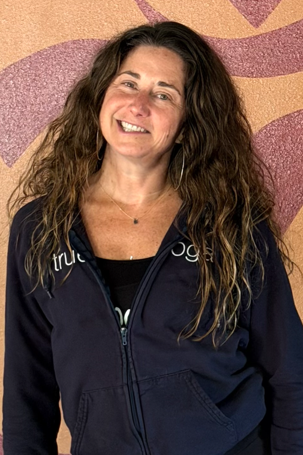 A smiling woman with long, curly brown hair, wearing hoop earrings, a black shirt, and a navy blue zip-up jacket with partial white text, standing in front of a pink and beige textured wall.
