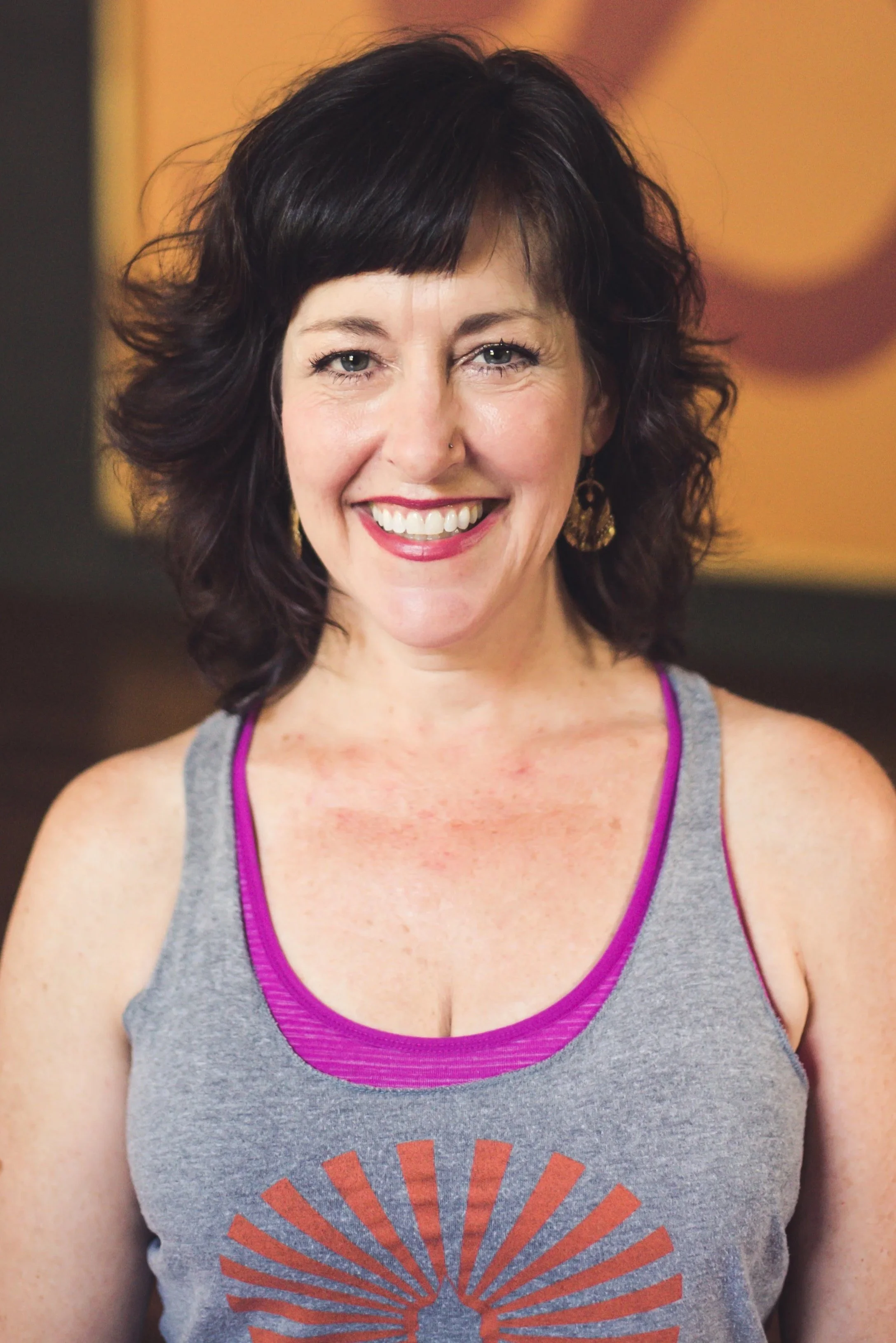 Close-up of a smiling woman with short dark curly hair, wearing a gray athletic tank top with purple straps and a sunburst logo on the front, and gold earrings, against a blurred warm-toned background.