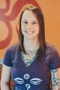 A young woman with red hair smiling, wearing a purple t-shirt with a graphic design, standing in front of a patterned orange background.
