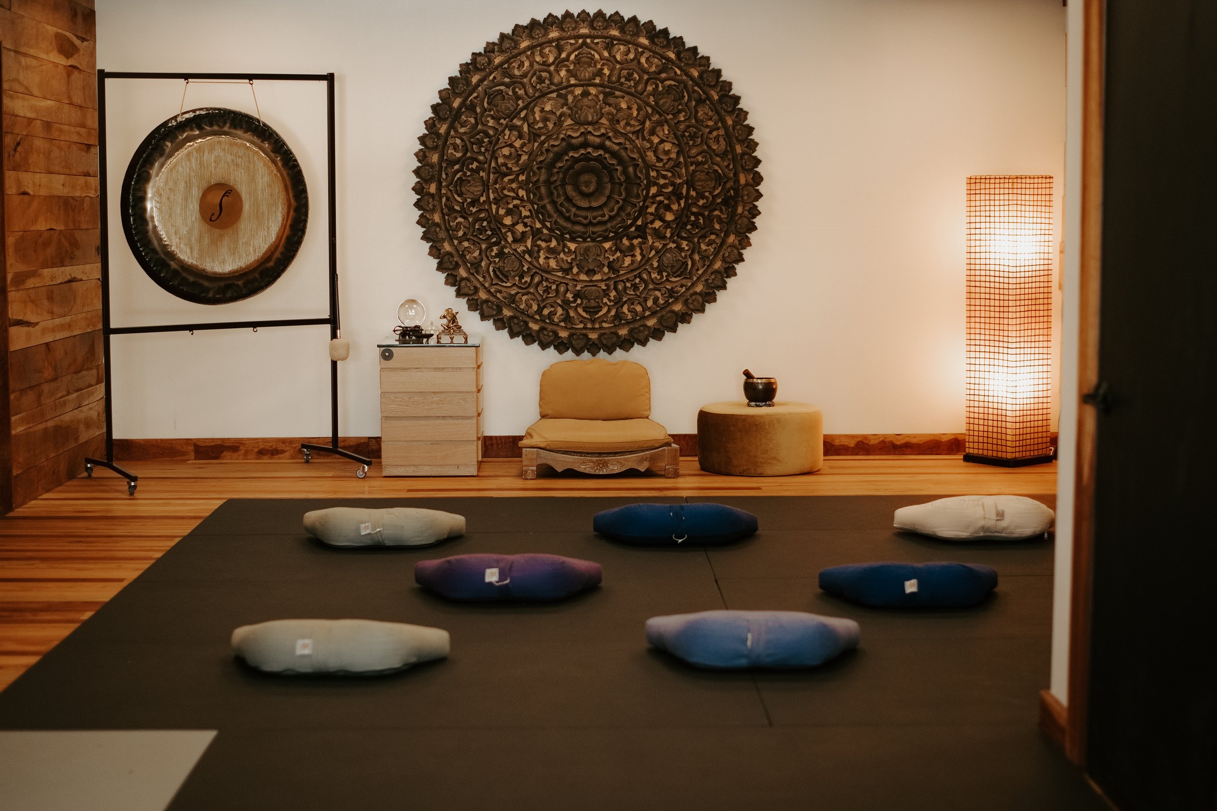 A meditation room with a black mat on the floor and round cushions in white, purple, and blue. The wall features a large, intricate wooden mandala art piece and a smaller wooden sculpture. To the right, there's a tall, grid-patterned floor lamp emitting warm light. The room has wood-paneled and white walls, creating a calming atmosphere.