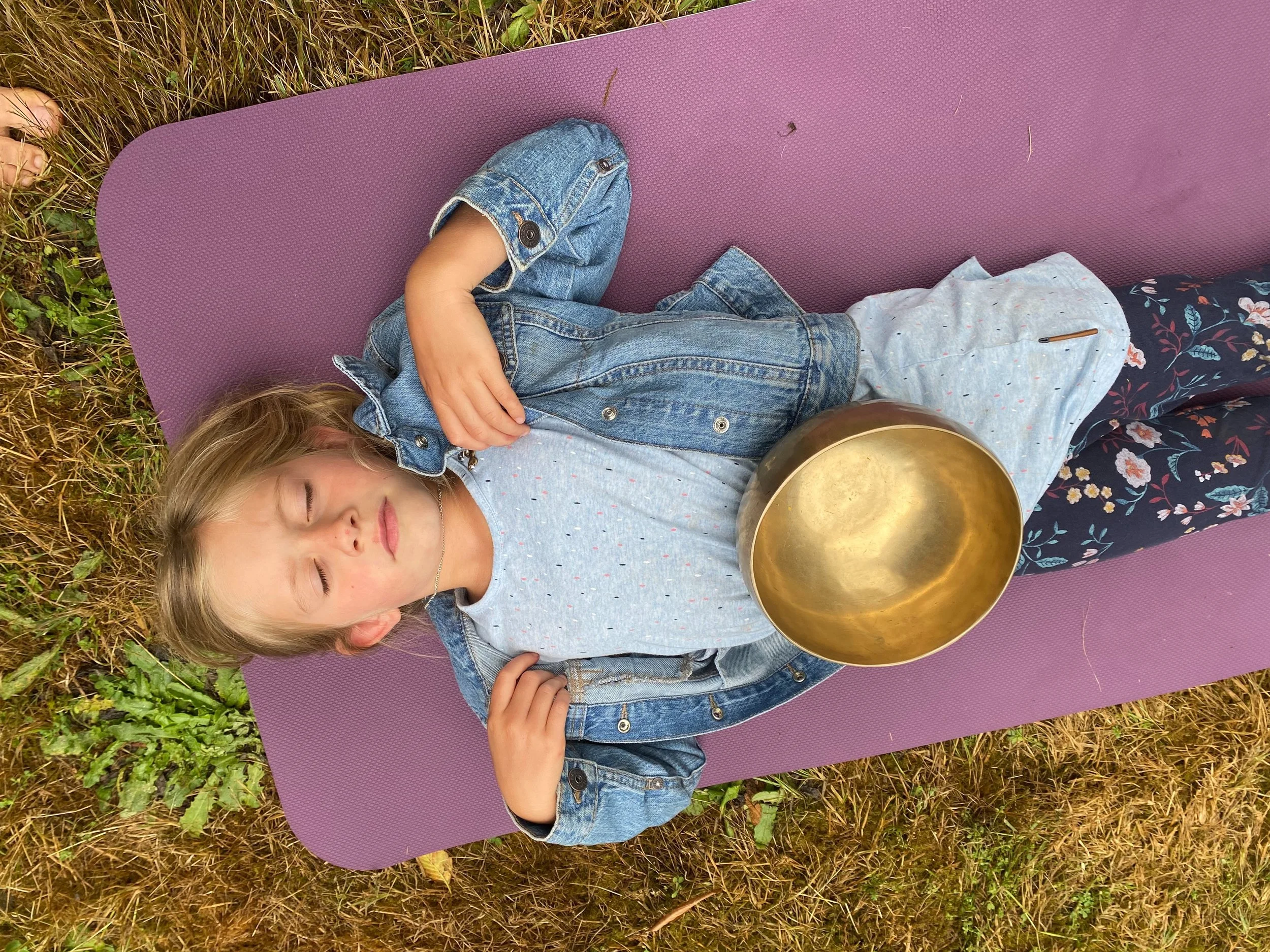 A young girl lying on a purple yoga mat outdoors with eyes closed, holding a brass singing bowl on her stomach, wearing a gray shirt, denim jacket, and floral-patterned pants.