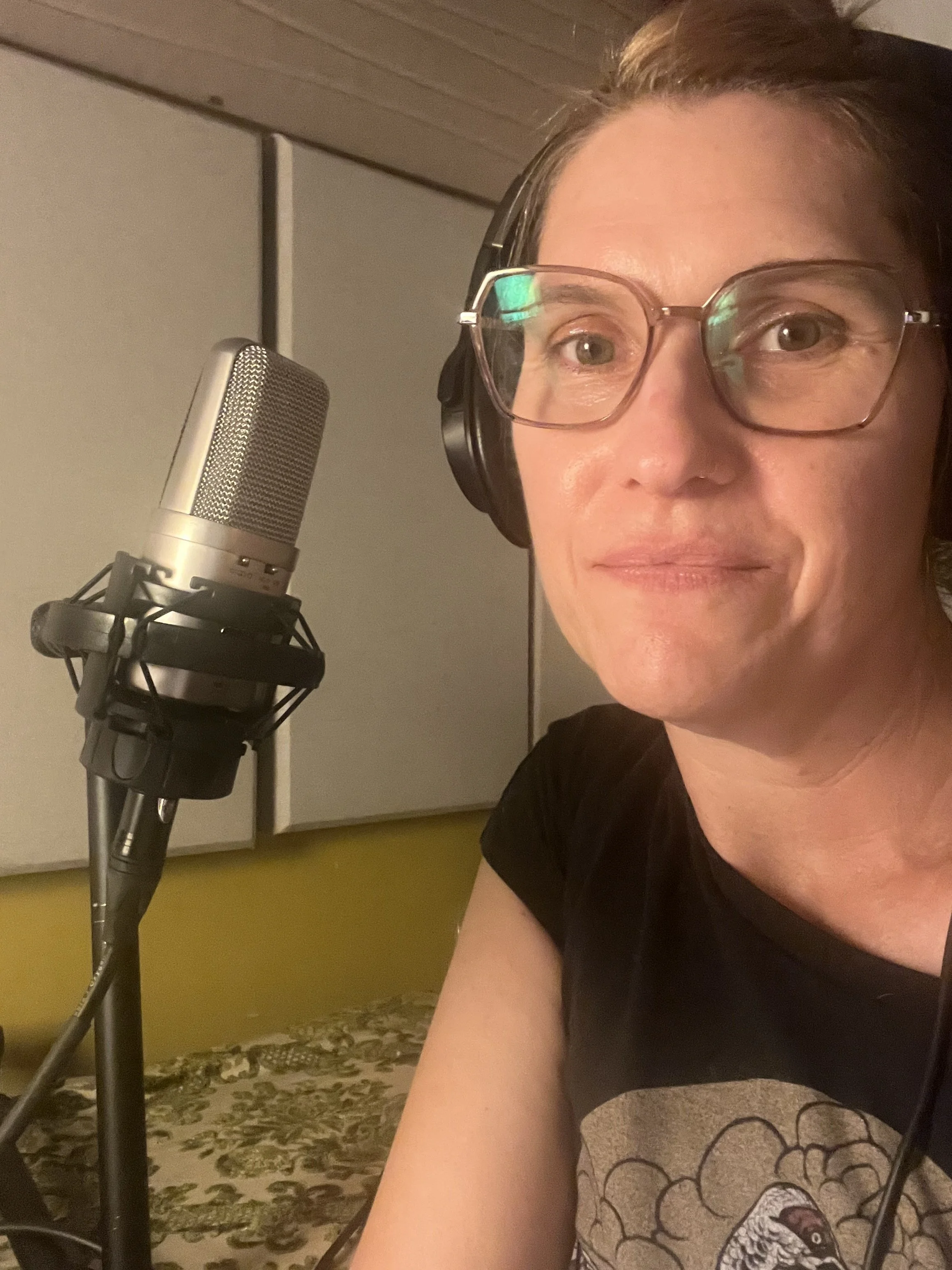 Cathy in her stdio, at the mic, wearing headphones, glasses, and a black t shirt