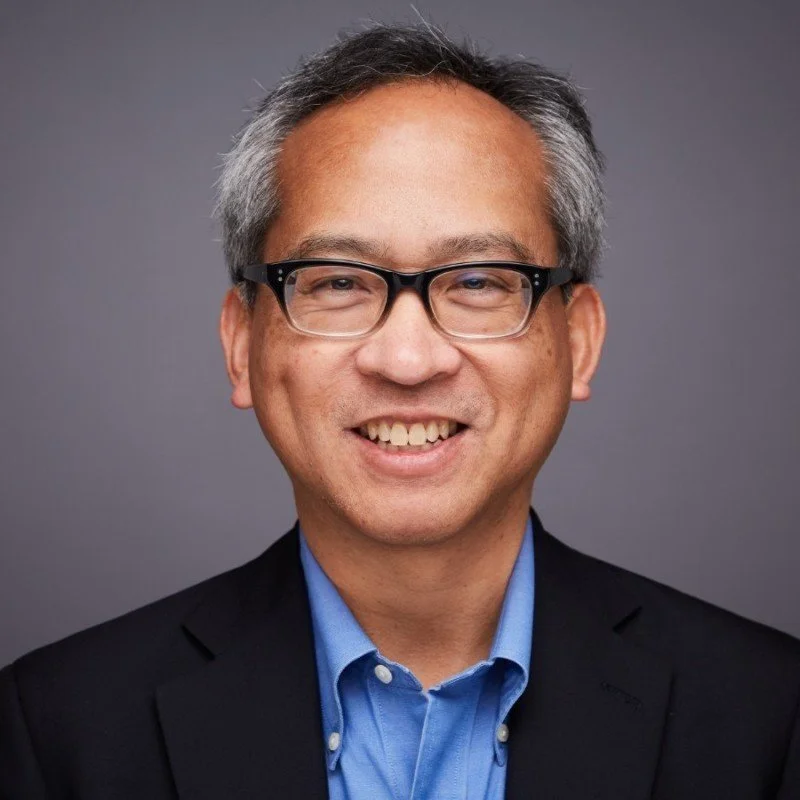 Professional headshot of a smiling Asian man with glasses, short gray hair, wearing a dark blazer and a blue collared shirt against a gray background.