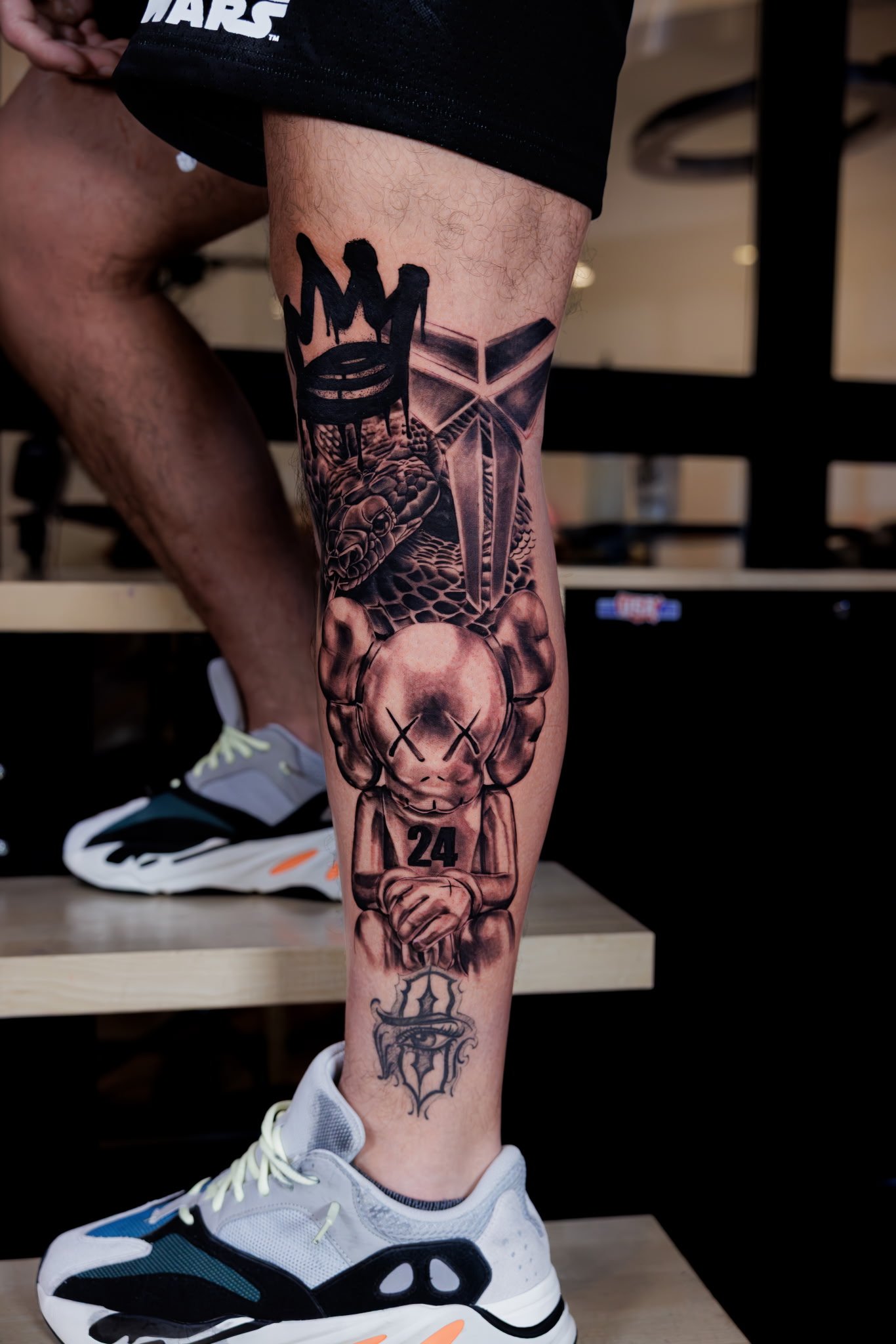 A person's leg with multiple tattoos, including a stylized heart with an X over it, a skull with snake details, and various other symbols, wearing white sneakers with black and teal accents.