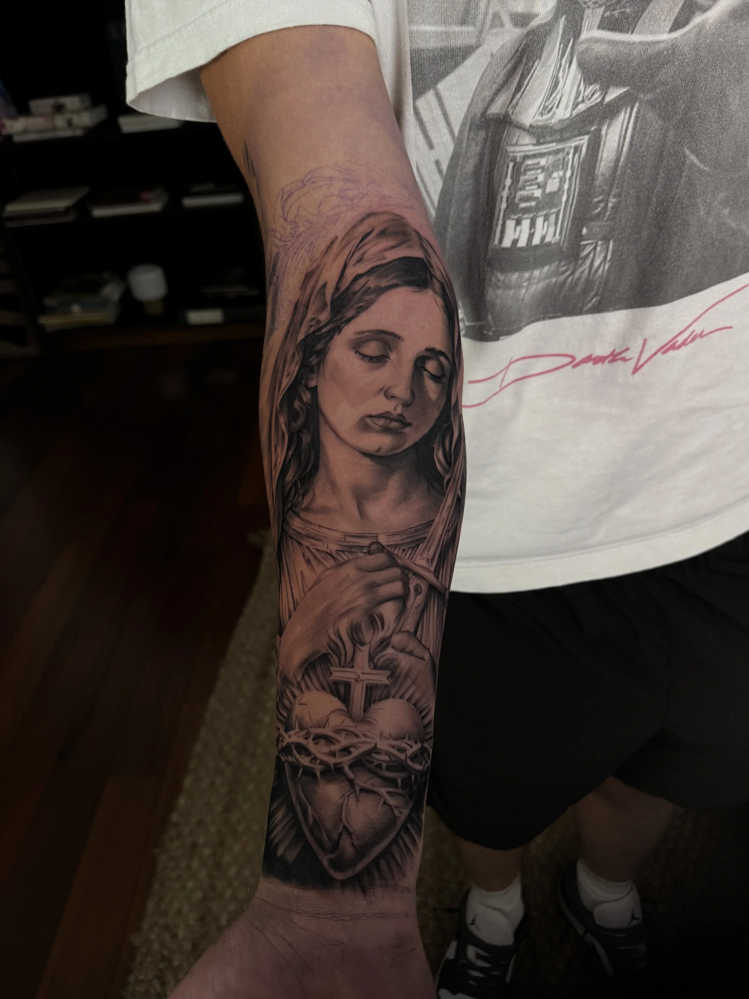 A detailed black and gray tattoo on a person's forearm depicting a woman with closed eyes and flowing hair, holding a large cross, with a sacred heart.