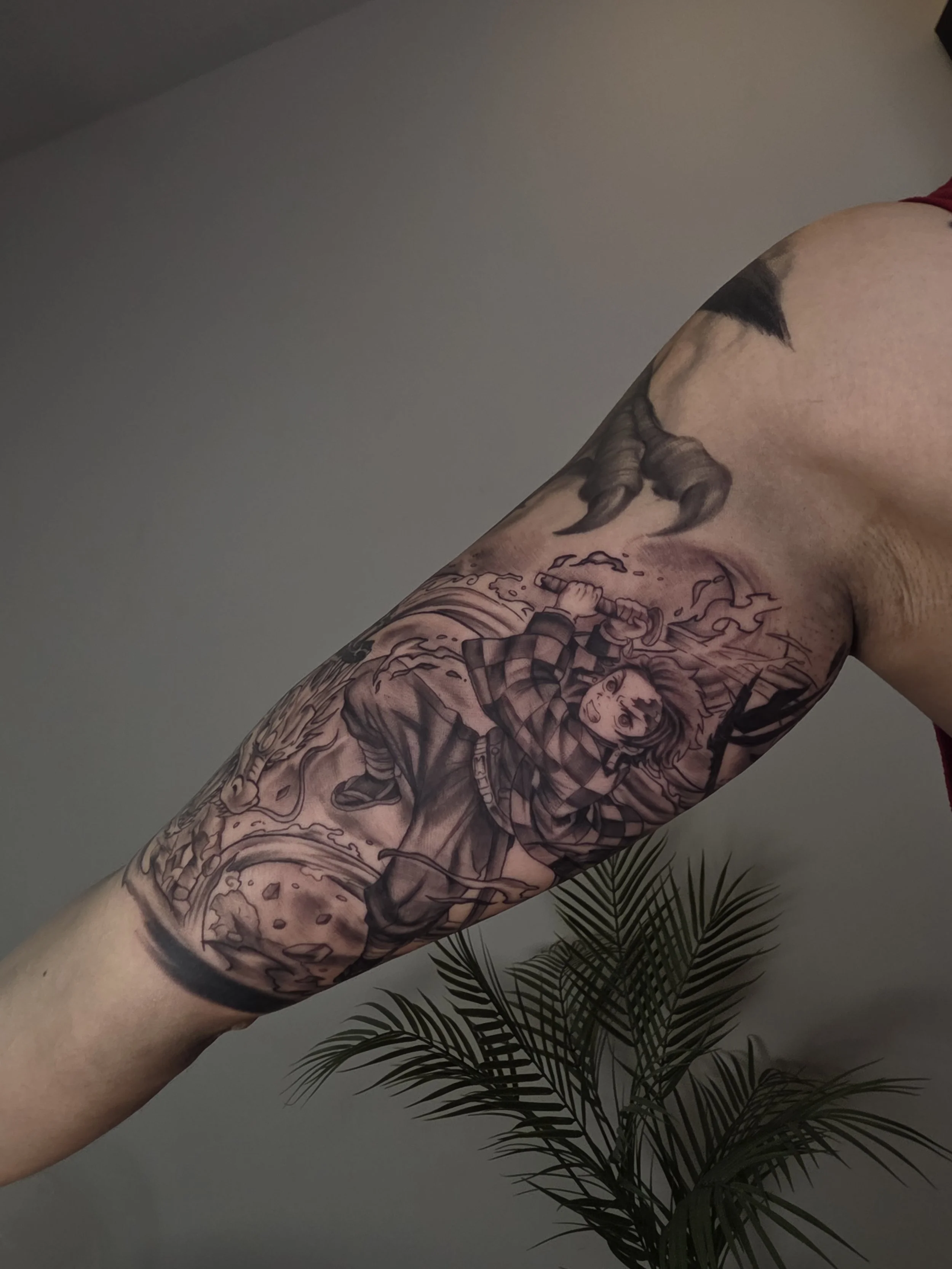 Close-up of a person's arm with a detailed black and gray tattoo sleeve featuring various characters, designs, and a wolf head. A plant with long, thin leaves is visible at the bottom.