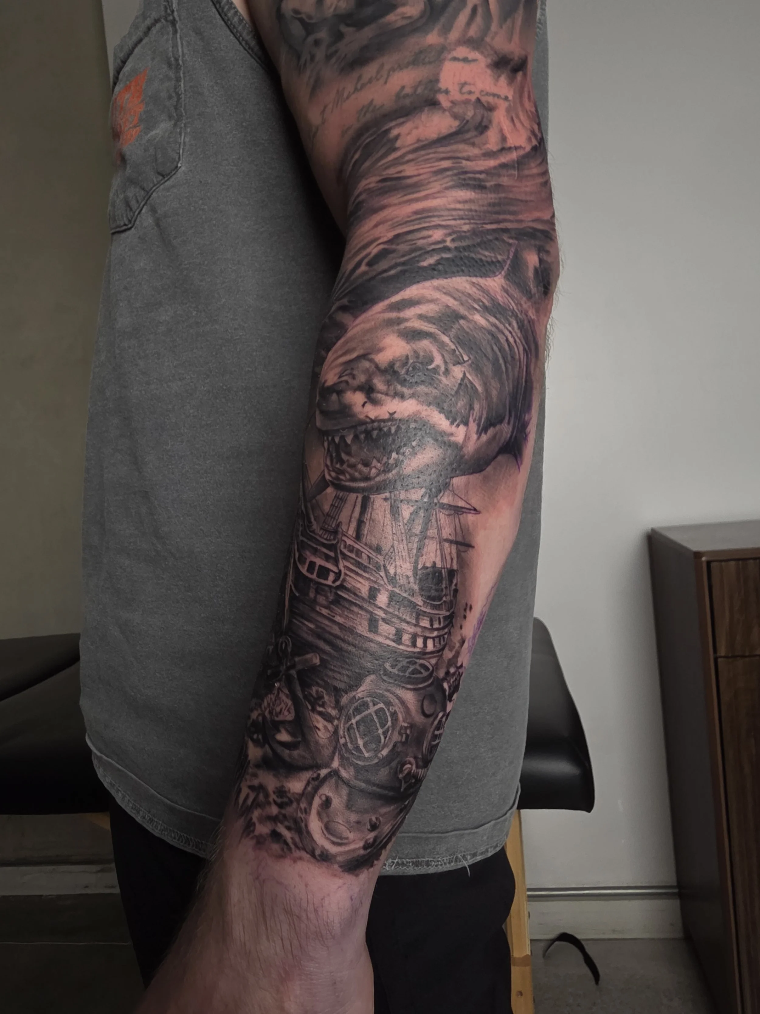 Close-up of a person's arm with a full sleeve tattoo depicting a wolf's head, ships, and ocean waves, with a person's upper body in the background wearing a gray t-shirt.