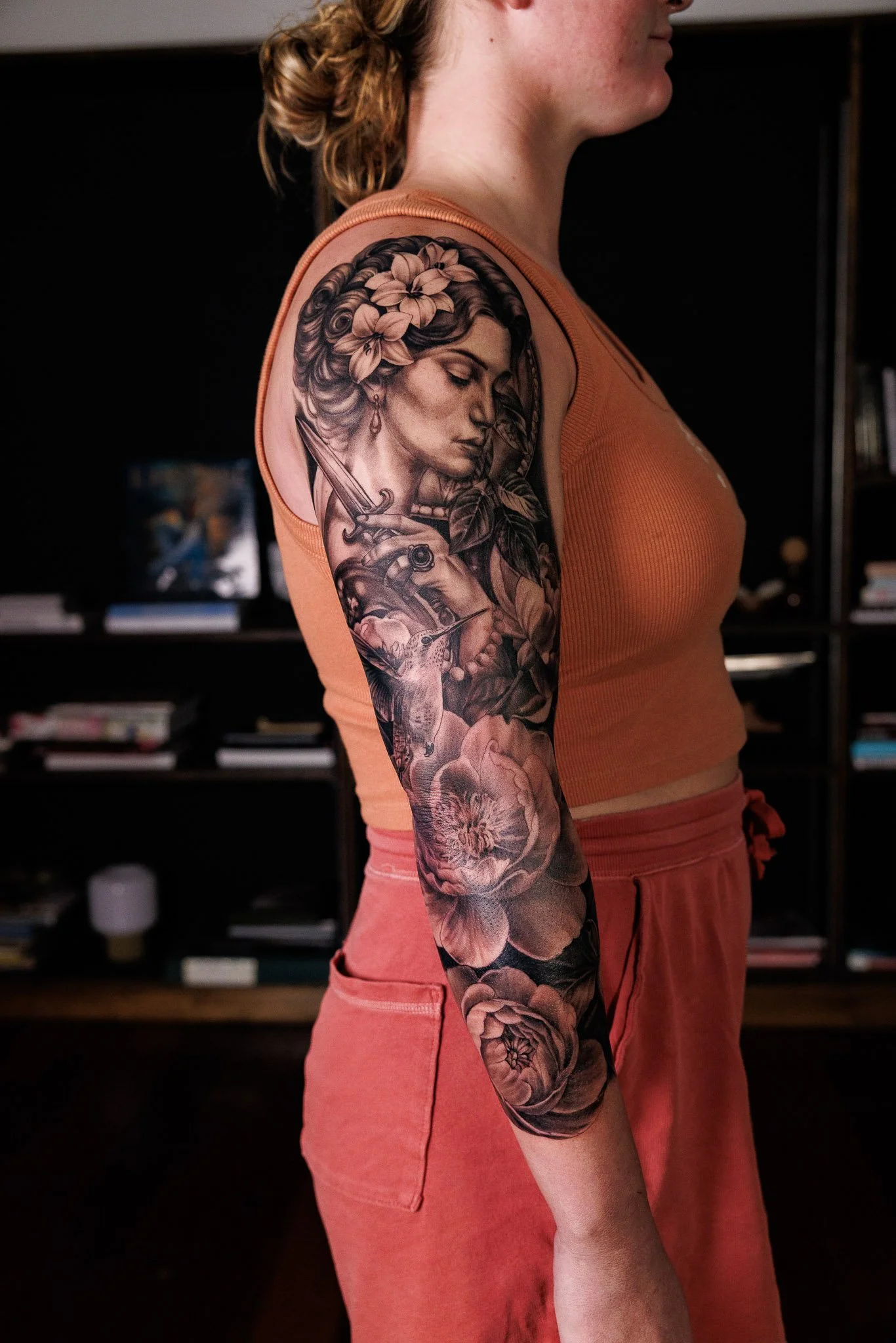 Close-up of a woman with a detailed black and gray tattoo sleeve on her left arm, featuring a woman's face with flowers in her hair and a dagger, wearing a sleeveless orange top and pink pants.