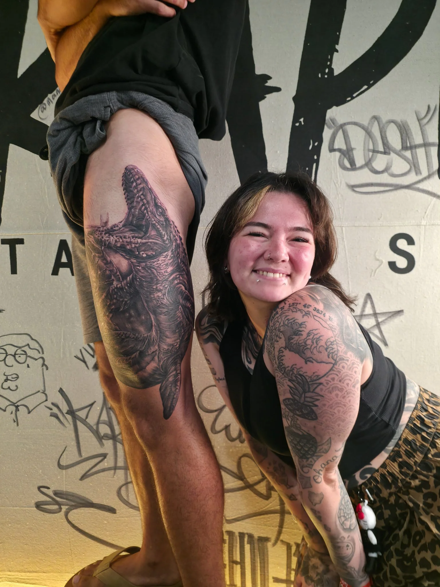A person with tattoos on their arm and shoulder, smiling and bending forward, with another person's leg visible in the background. The setting appears to be a tattoo studio with graffiti on the wall.