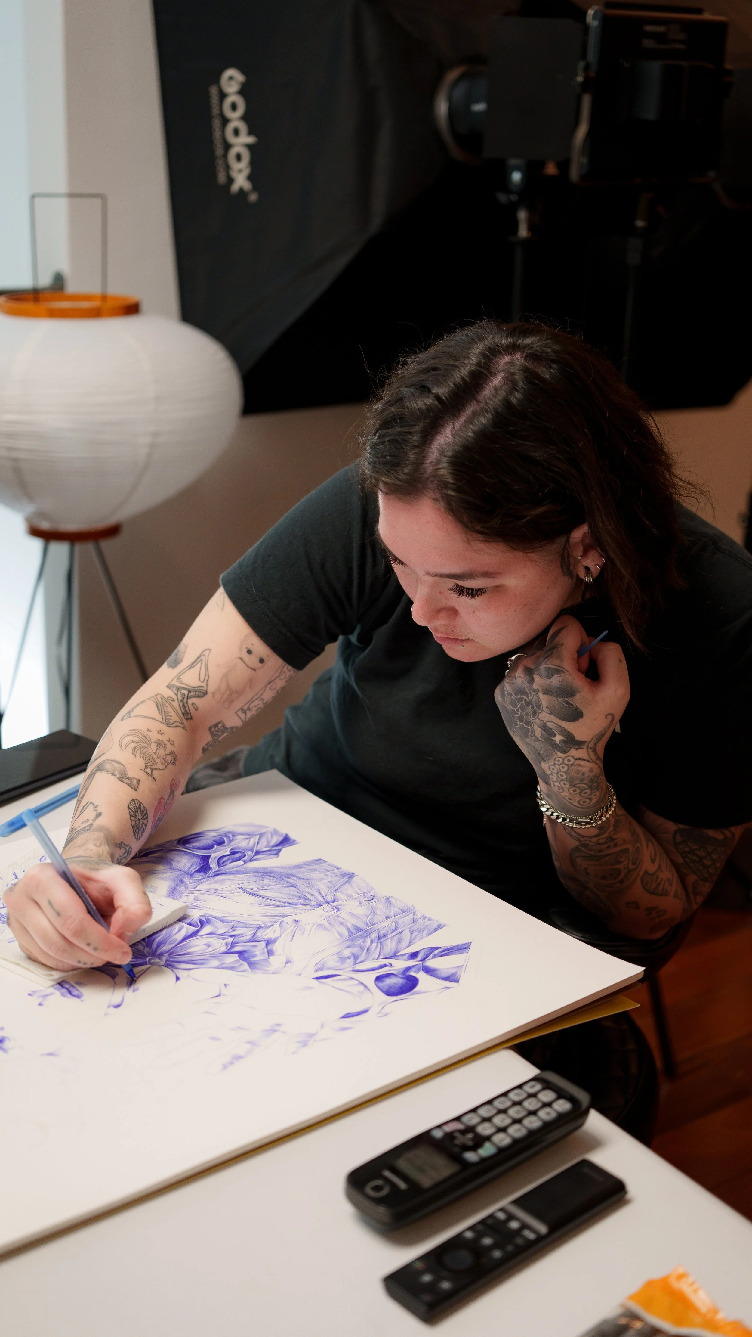 A woman with tattoos on her arms and dark hair is drawing a detailed purple illustration of a woman with roses on white paper at a desk. There are remote controls and a calculator on the desk, and a lamp to her left. A black backdrop with the brand name 'Godox' is visible behind her.