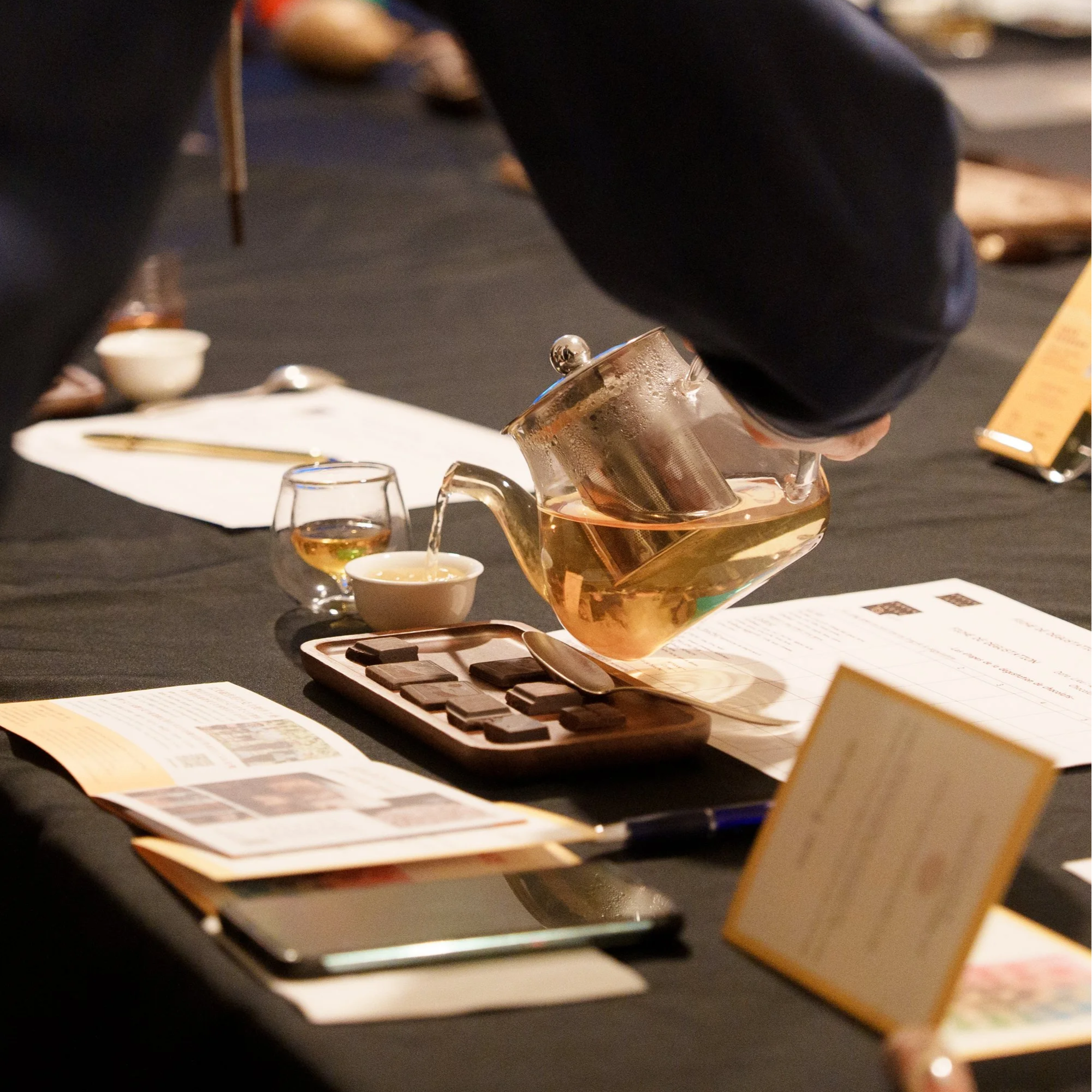 Une table avec des notes de la dégustation, des tasses contenant d'une infusion de cacao, des morceaux de chocolat et une main versant une infusion de cacao.