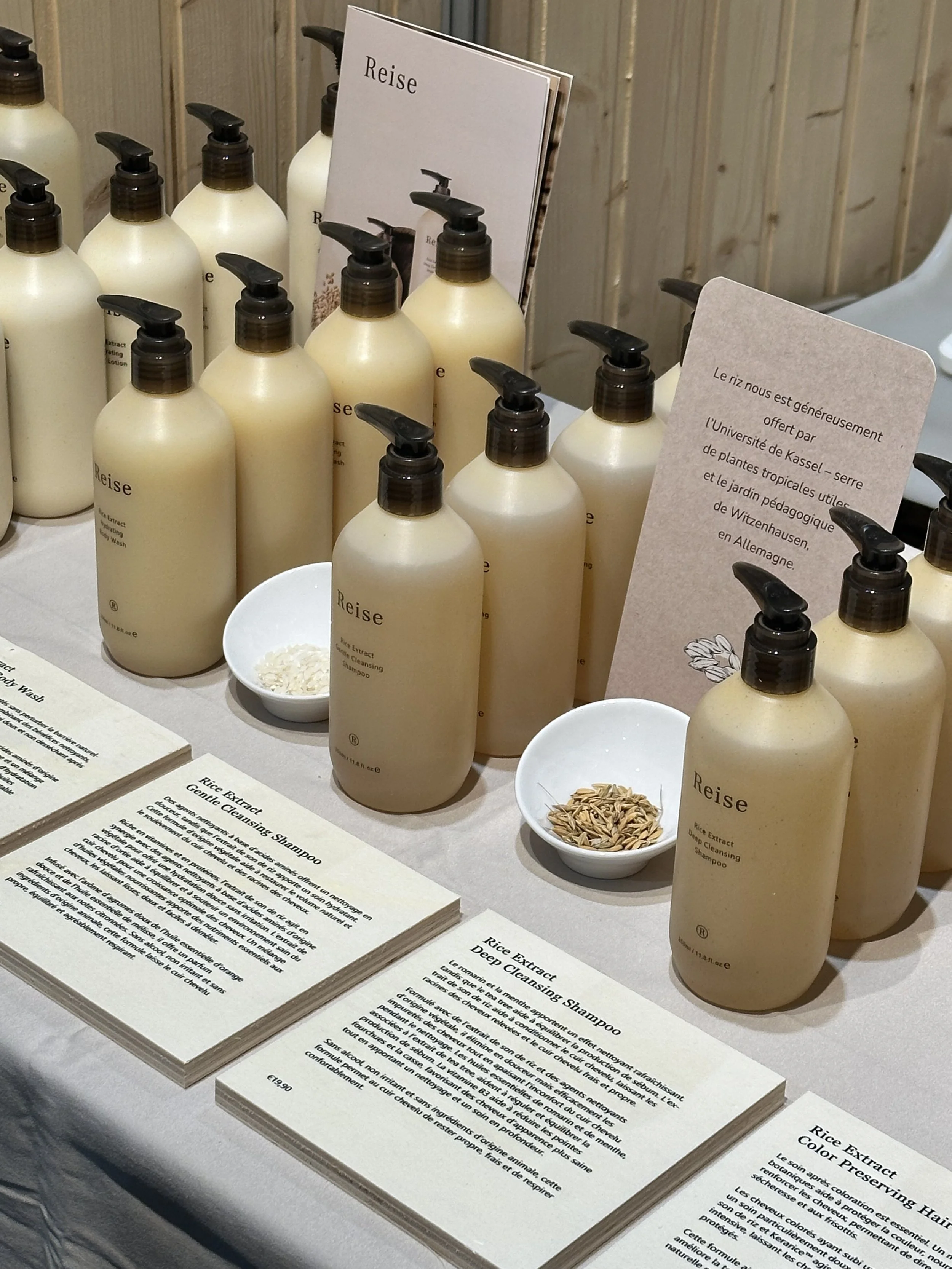 Échantillons de bouteilles beige de shampooing et savons à pompe sur une table dans un salon avec des panneaux d'informations écrits en français