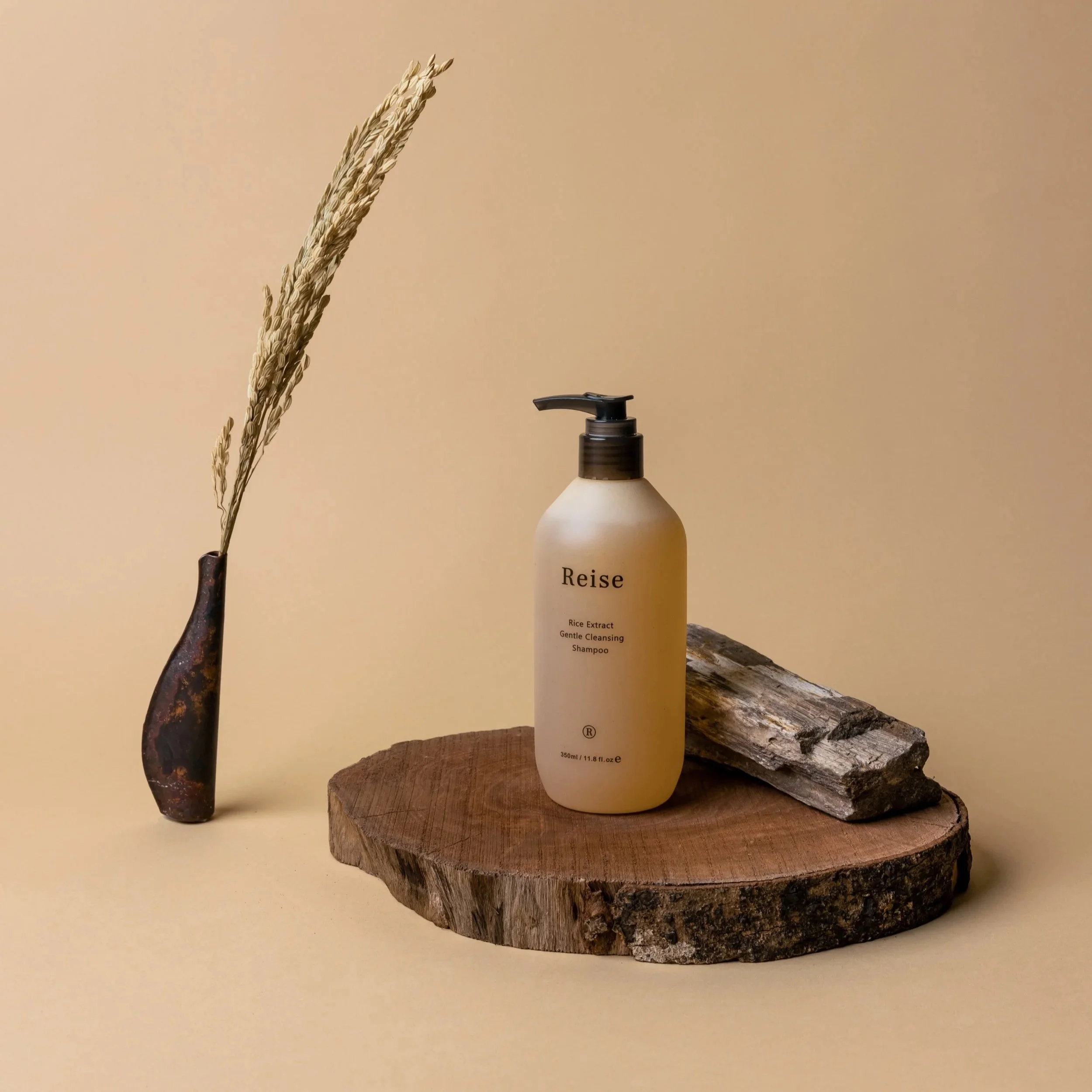 Une bouteille de shampoing au nom 'Reise' place sur un morceau de bois, avec une branche d'herbe sèche à gauche et une branche de bois à droite, le tout sur un fond beige.